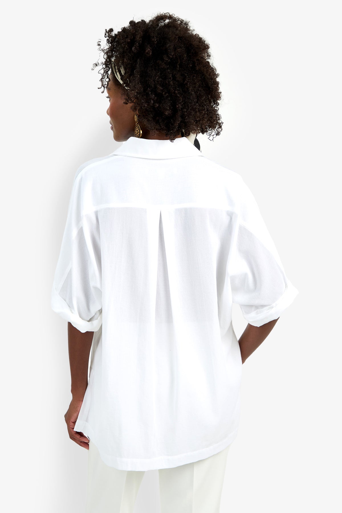 A woman with curly hair wears the Hawaii Top, a loose-fitting white short-sleeve blouse, paired with white pants. She faces away from the camera to display the back details of the blouse against a plain white background.