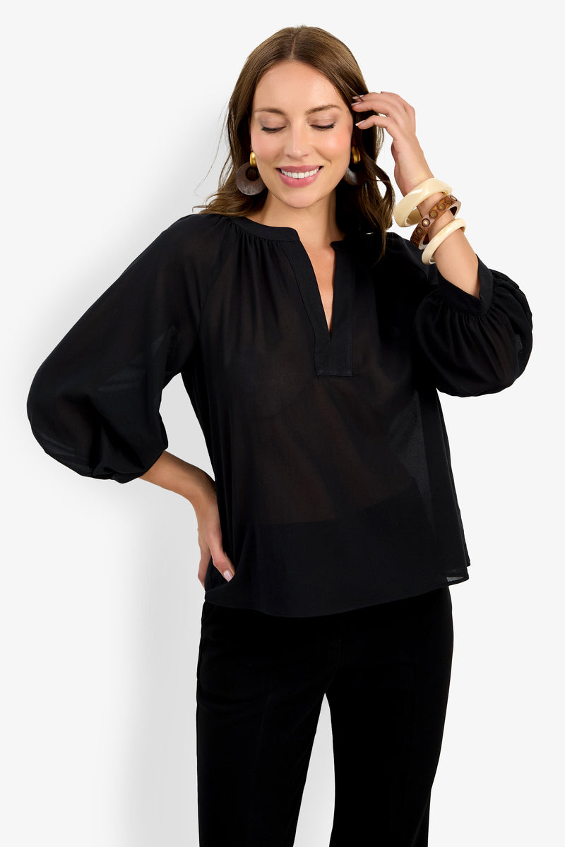 Smiling woman in the Light Hearted Top, a loose black long-sleeve V-neck blouse, paired with black pants and chunky bracelets, standing with one hand on her hip and the other touching her hair.