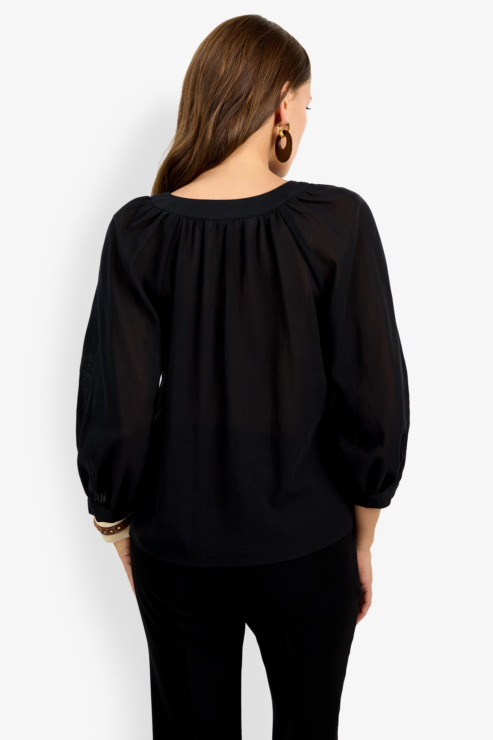 A woman with long brown hair is seen from the back wearing the Light Hearted Top, a loose black blouse with gathered details and balloon sleeves, paired with black pants. She accessorizes with hoop earrings and bracelets.