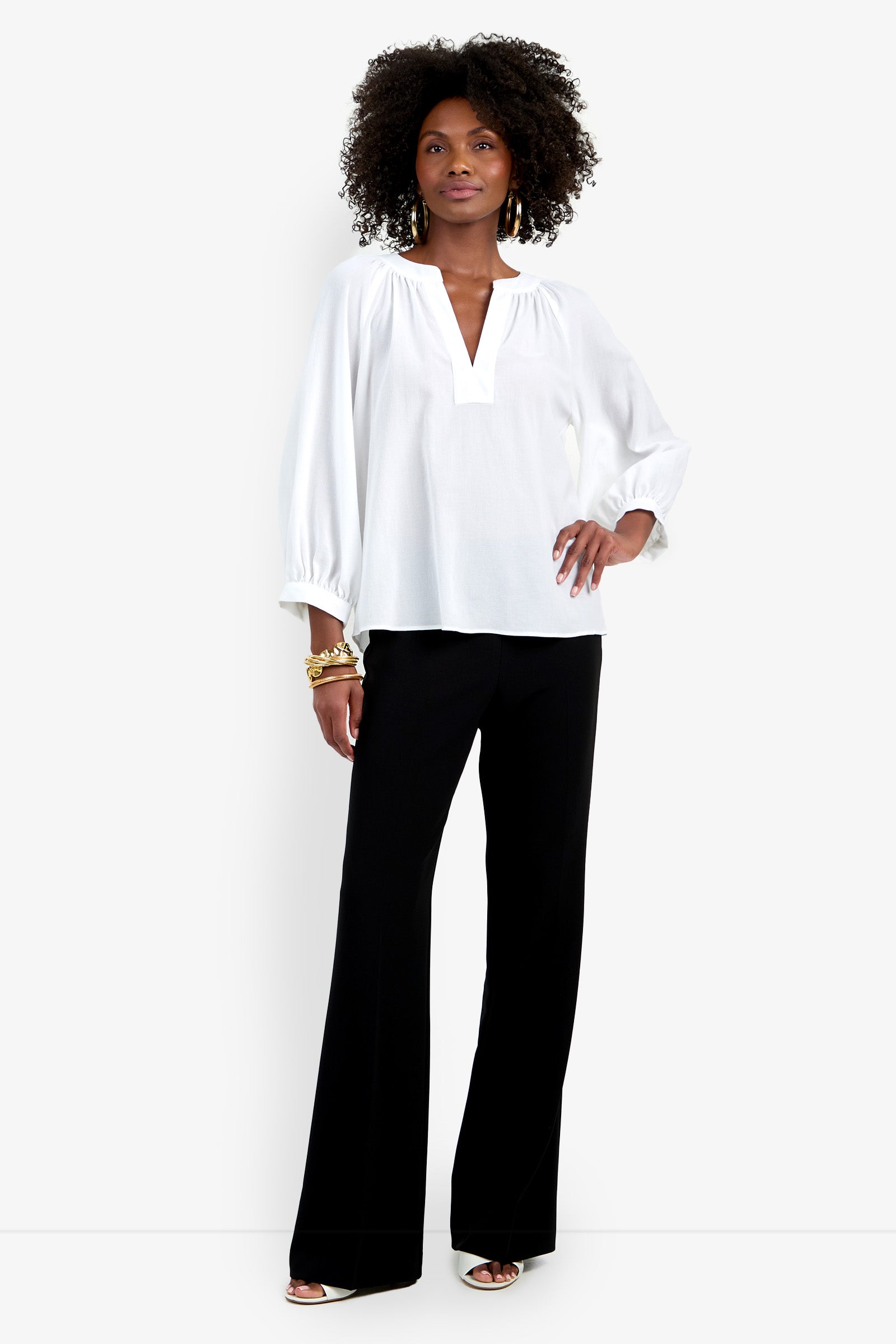 A woman stands confidently wearing the Light Hearted Top, a loose white V-neck blouse with long sleeves, paired with black wide-leg trousers, open-toed heels, gold bracelets, and earrings.