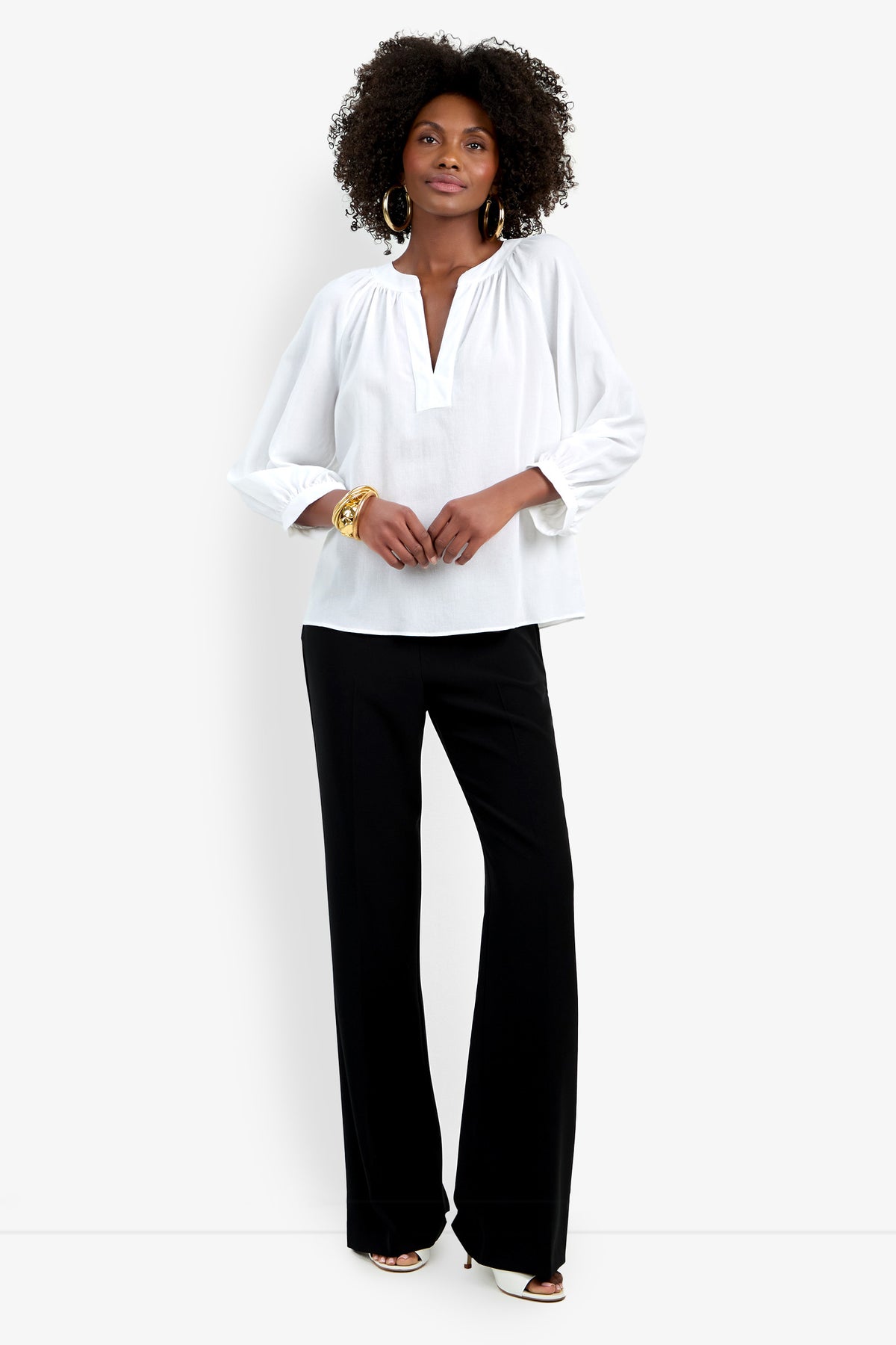 A woman stands against a white background wearing the Light Hearted Top, a loose white long-sleeve V-neck blouse, paired with black wide-leg pants. She completes the look with hoop earrings, bracelets, and white shoes.