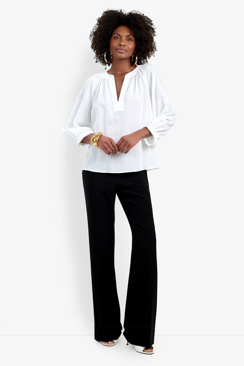 A woman stands against a white background wearing the Light Hearted Top, a loose white long-sleeve V-neck blouse, paired with black wide-leg pants. She completes the look with hoop earrings, bracelets, and white shoes.