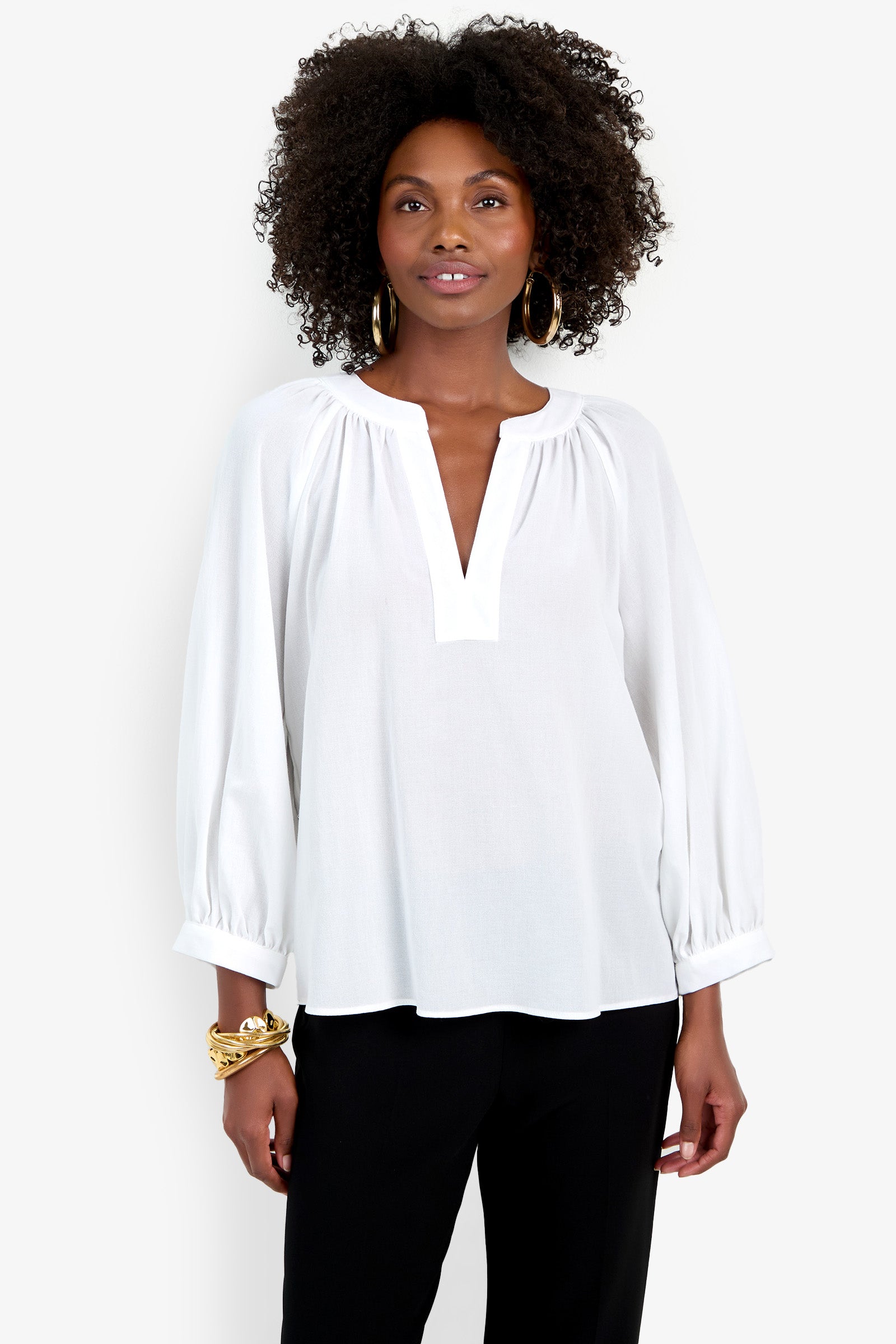 A woman with curly hair wears the Light Hearted Top, a white long-sleeve blouse with a V-notch neckline, paired with black pants. She accessorizes with hoop earrings and gold bracelets, standing against a plain white background.