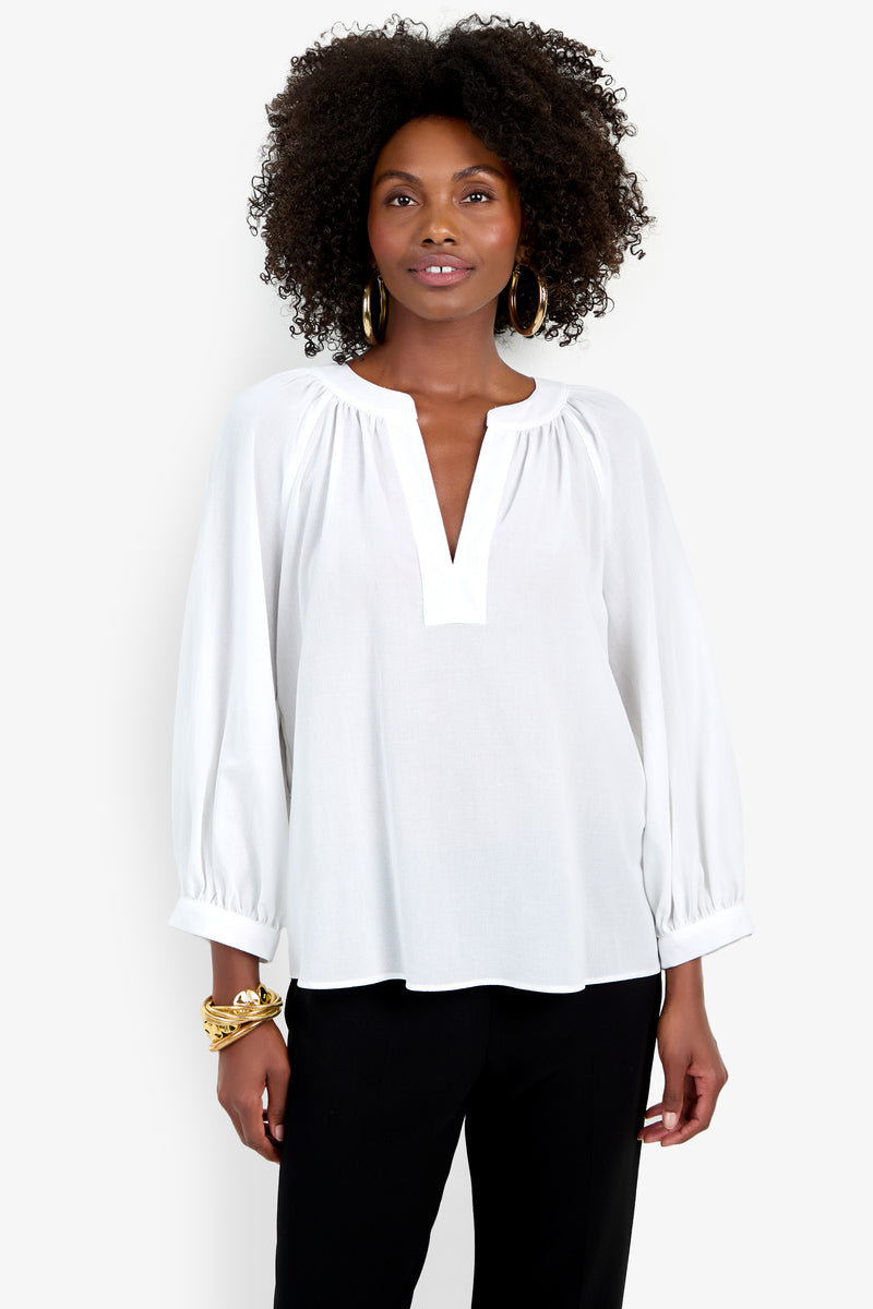 A woman with curly hair wears the Light Hearted Top, a white long-sleeve blouse with a V-notch neckline, paired with black pants. She accessorizes with hoop earrings and gold bracelets, standing against a plain white background.