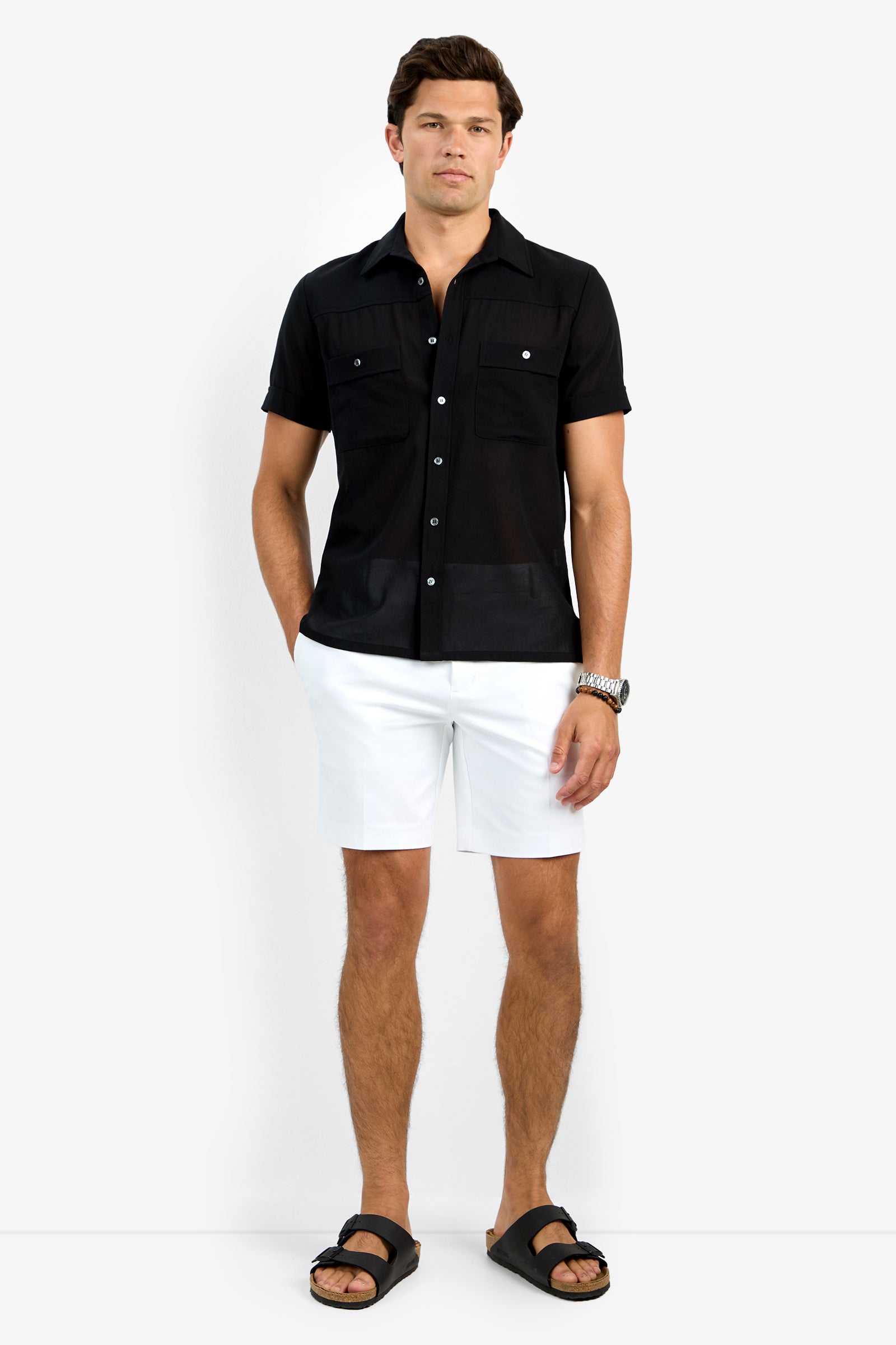 A man stands against a plain white background wearing the Will Shirt, a short-sleeve black button-up, paired with white shorts, black sandals, and a wristwatch.