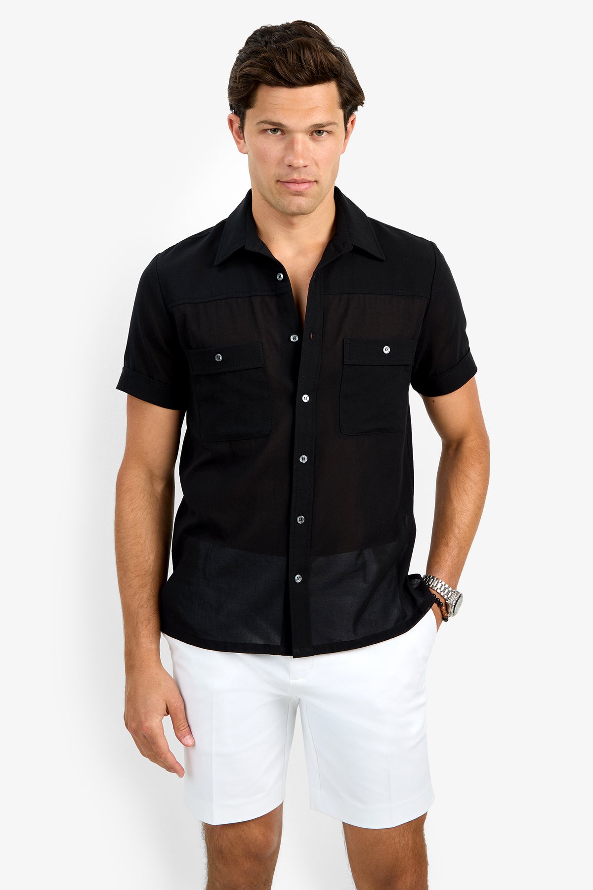 A man wears the Will Shirt, a short-sleeve black button-up with front pockets, and white shorts while standing against a plain white background. He looks straight ahead with one hand in his pocket and the other by his side.