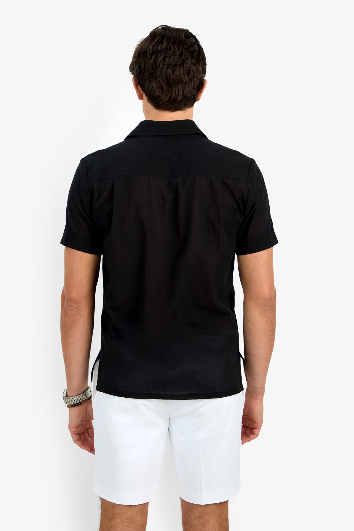 A man with short dark hair wears the Will Shirt, a short-sleeved black button-up, paired with white shorts and a watch. He stands facing away on a plain white background.