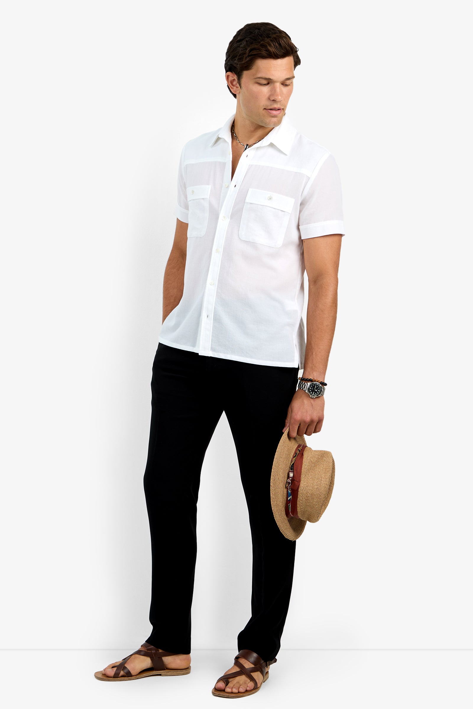 A man stands facing slightly to the side, wearing the Will Shirt—a white short-sleeve button-up—paired with black pants, brown sandals, and holding a straw hat with a brown band in his left hand.