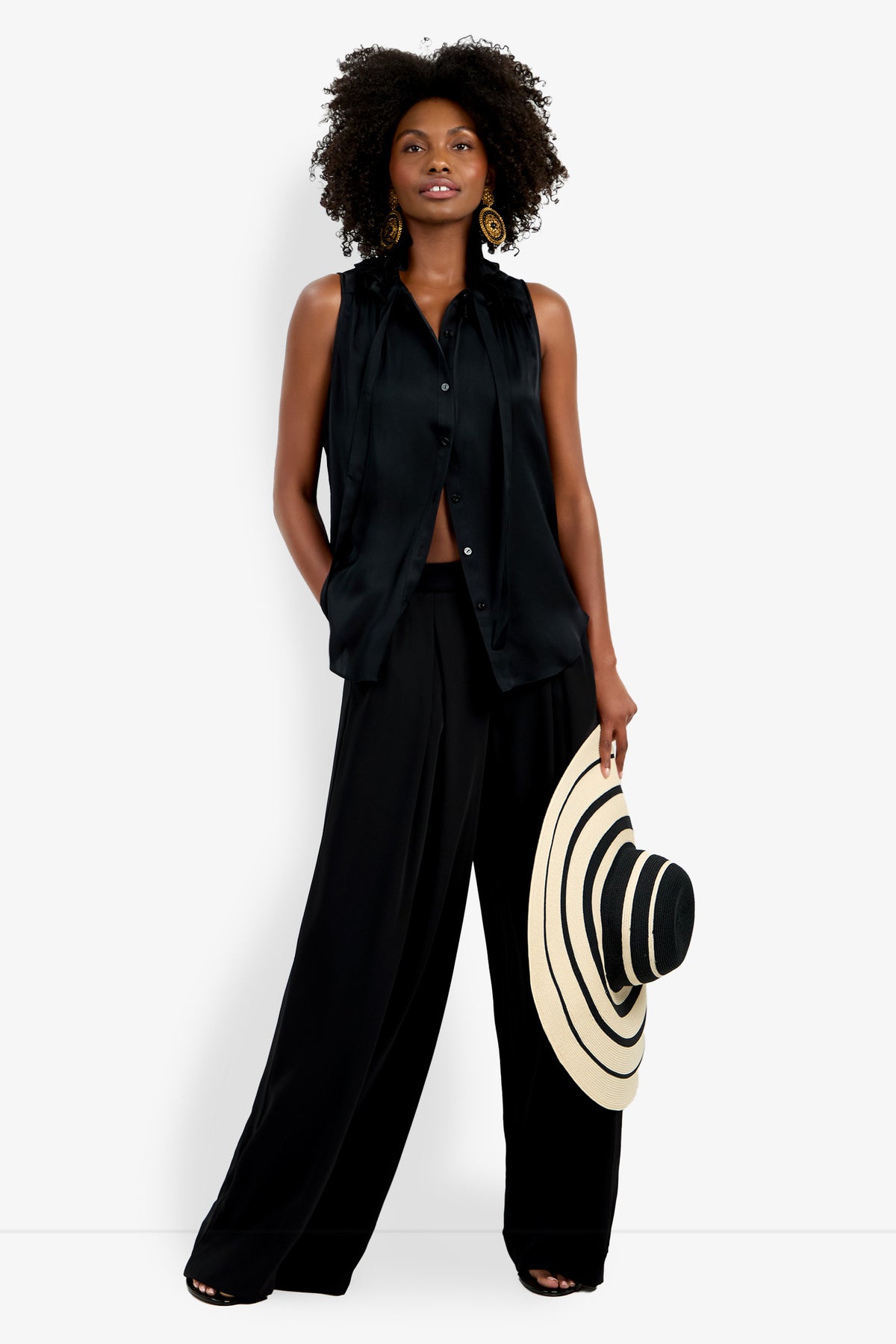 Wearing the Cinzia Top, a sleeveless black button-up blouse, and wide-leg black pants, a woman holds a wide-brimmed black-and-beige striped hat, standing confidently against a plain white background.