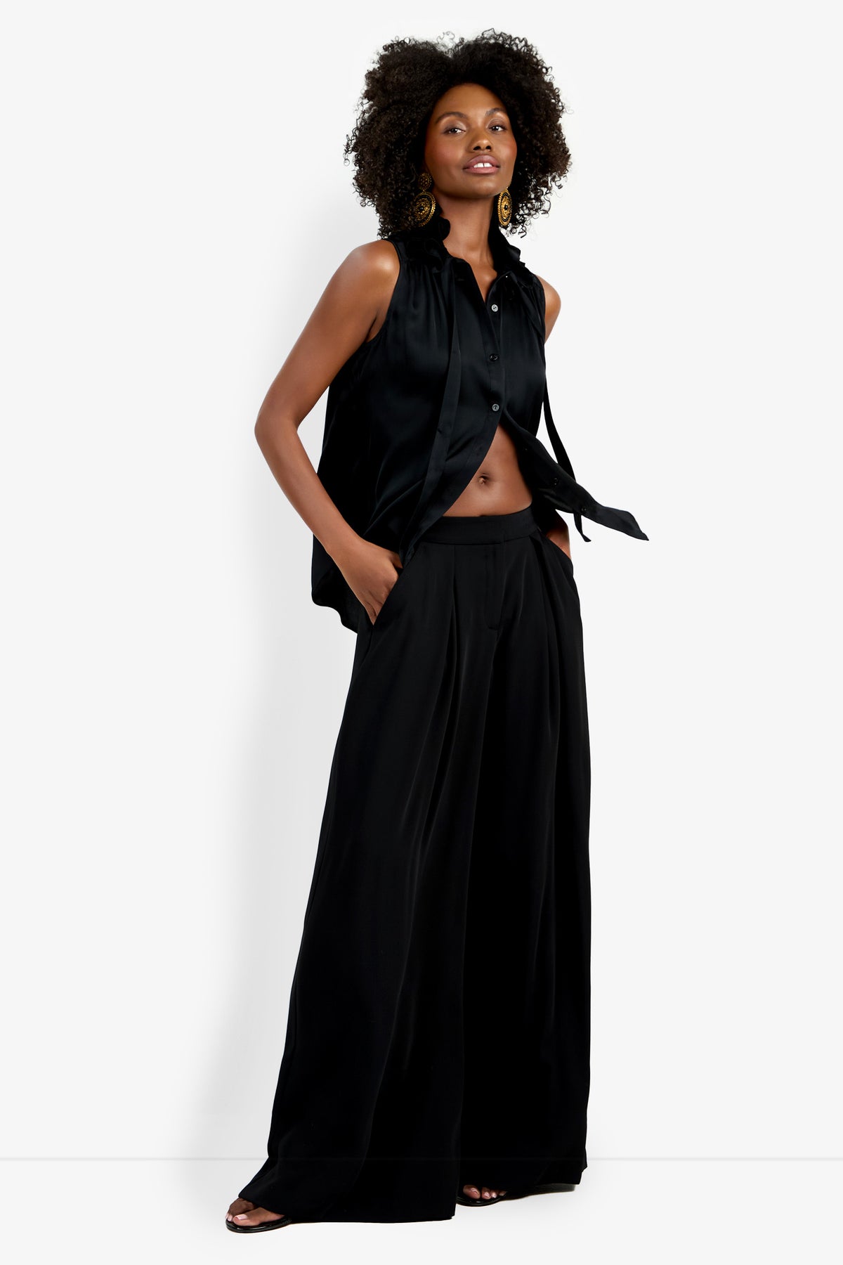 A woman stands confidently with one hand in her pocket, wearing the sleeveless black Cinzia Top and wide-leg black pants. Her hair is styled in natural curls as she poses against a white background.