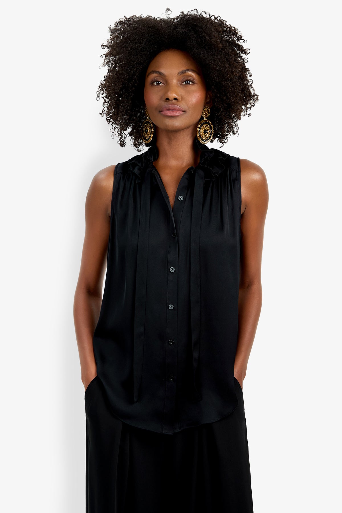 A woman with curly hair wears large gold earrings and the Cinzia Top, a sleeveless black button-down blouse with a tie neckline, standing with her hands in her pockets against a plain white background.