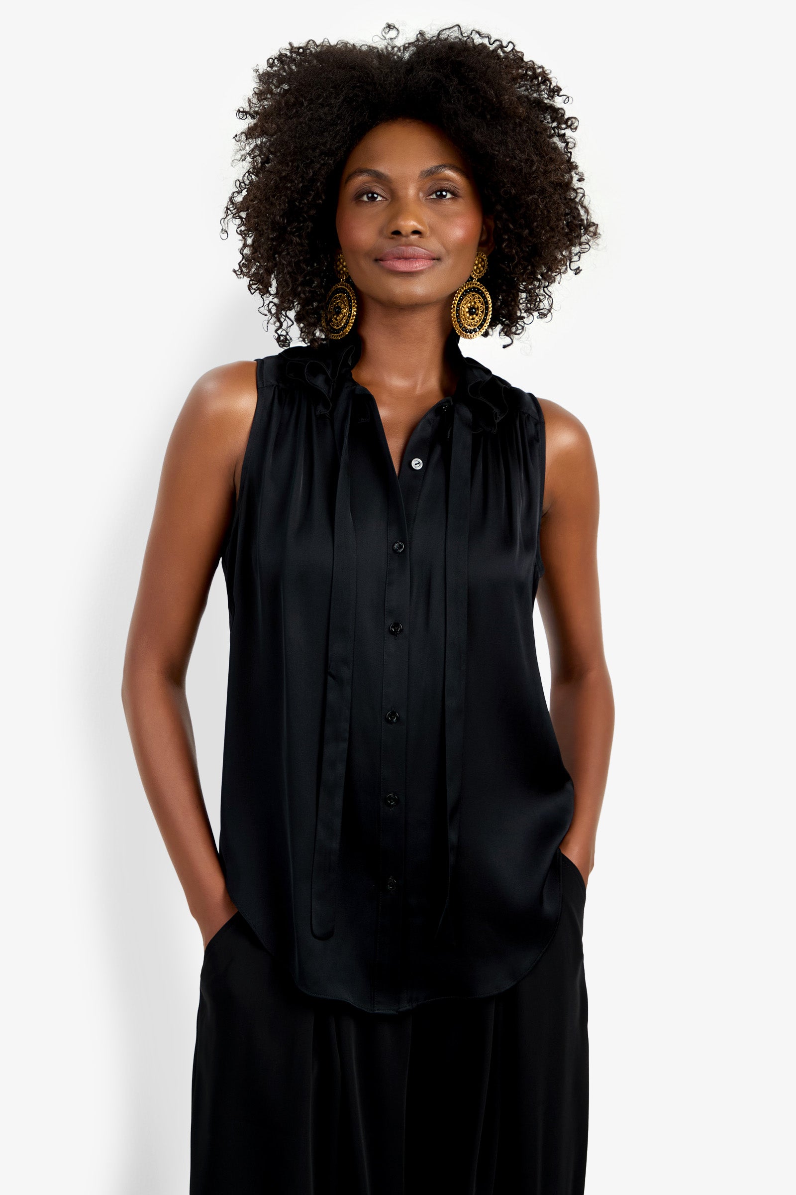 A woman with curly hair, large gold earrings, and hands in her pockets models the Cinzia Top, a loose-fit sleeveless black button-up blouse, against a plain white background.