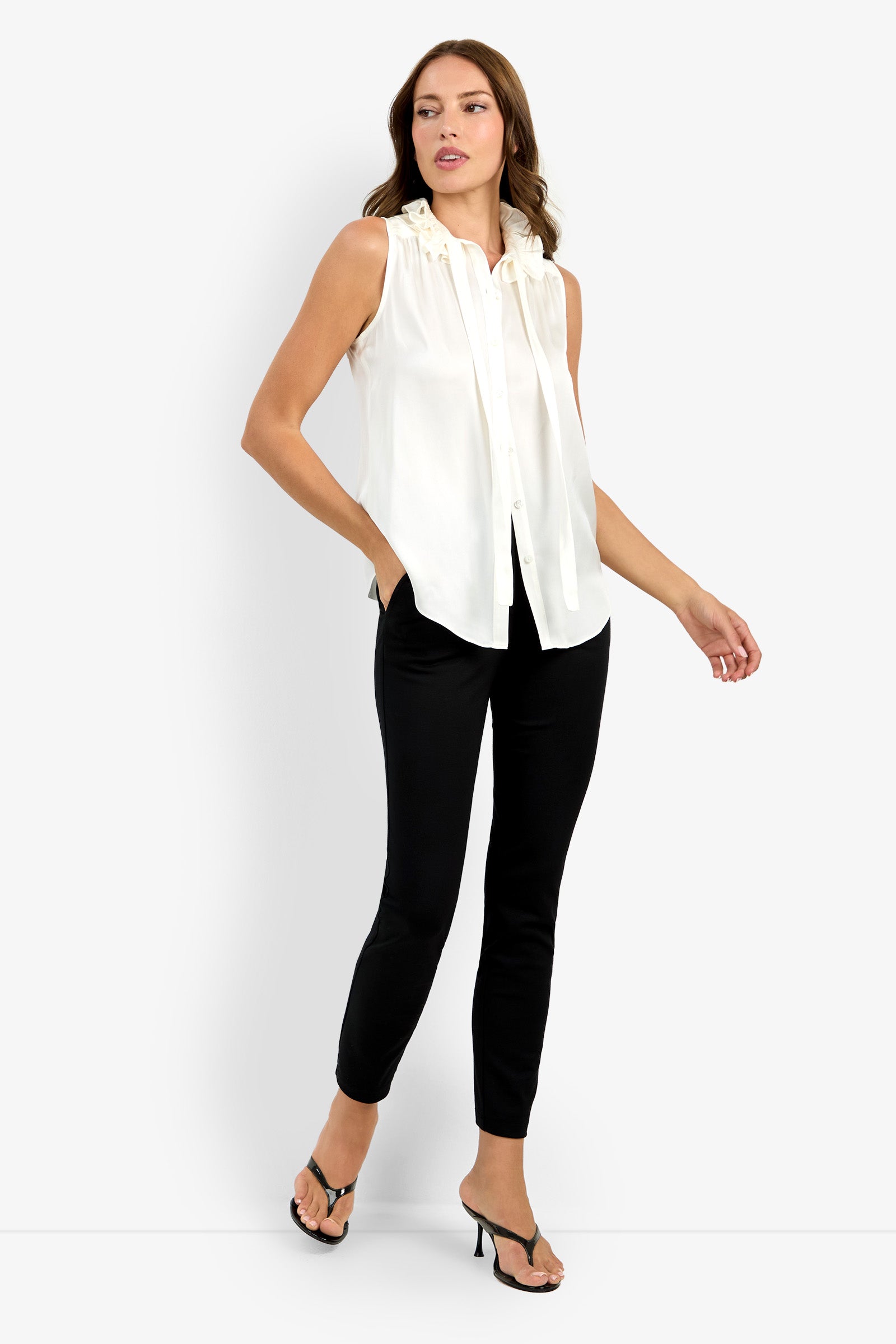 A woman with light brown hair poses against a white background, wearing the sleeveless Cinzia Top featuring a neck bow, paired with black slim-fit pants and black heeled sandals. One hand is slightly raised.