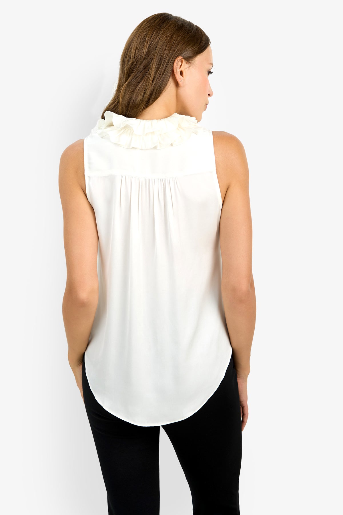 A woman with brown hair is seen from behind wearing the Cinzia Top, a sleeveless white blouse featuring ruffle details on the collar, paired with black pants, standing against a plain white background.