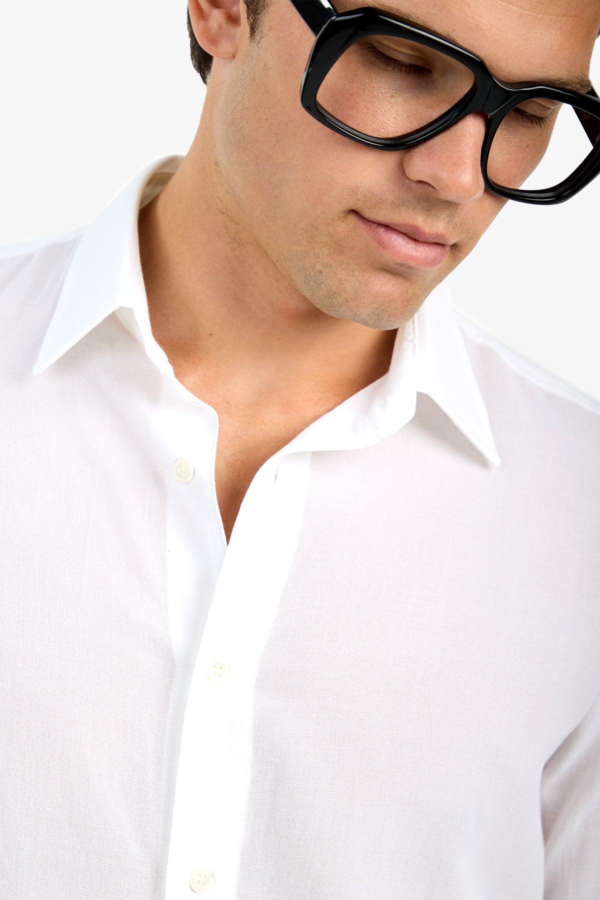 A person wearing the Aaron Shirt, a white collared shirt, along with large black glasses is shown from the shoulders up, looking down with a neutral expression against a plain background.
