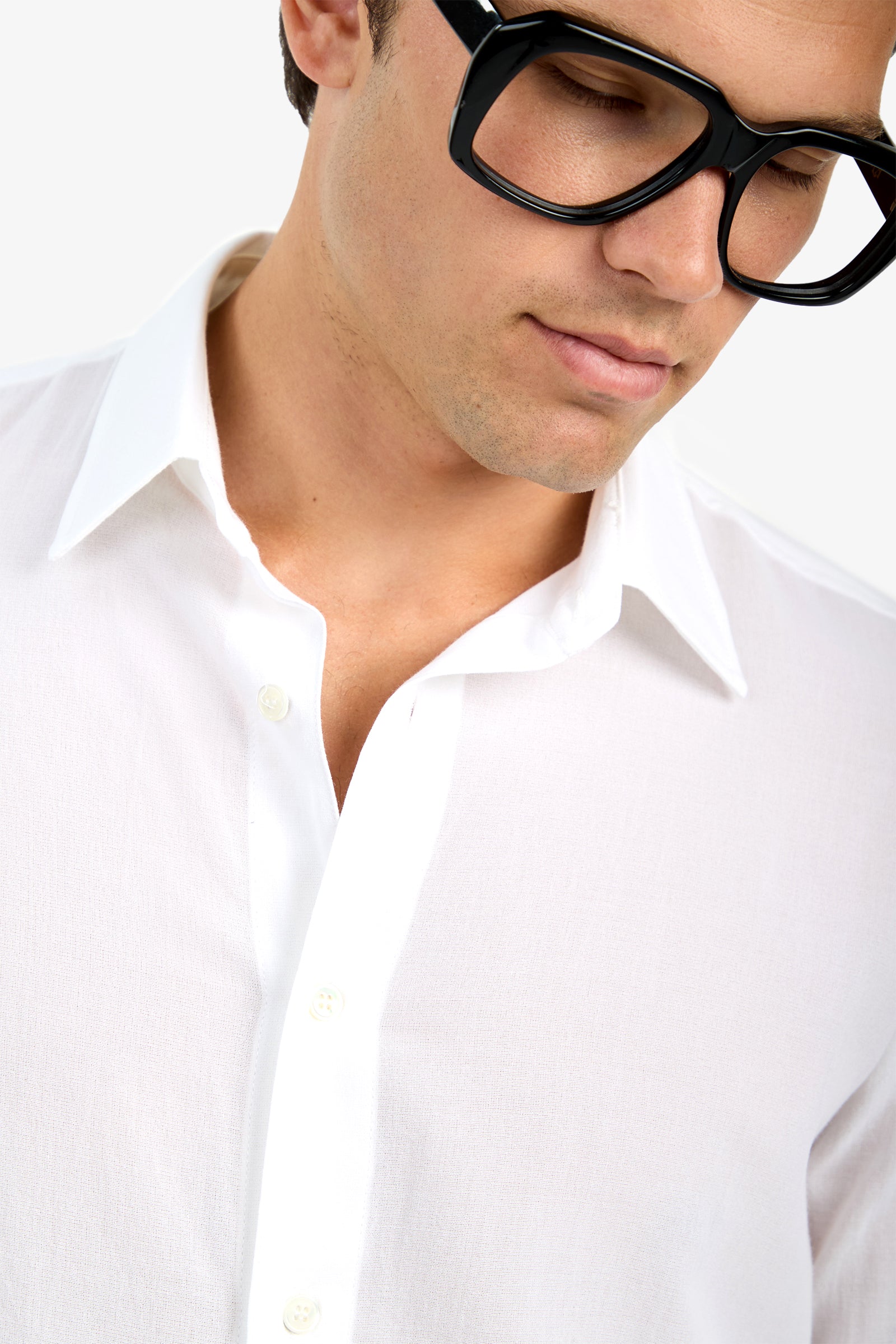 A person wearing the Aaron Shirt, a white collared shirt, along with large black glasses is shown from the shoulders up, looking down with a neutral expression against a plain background.