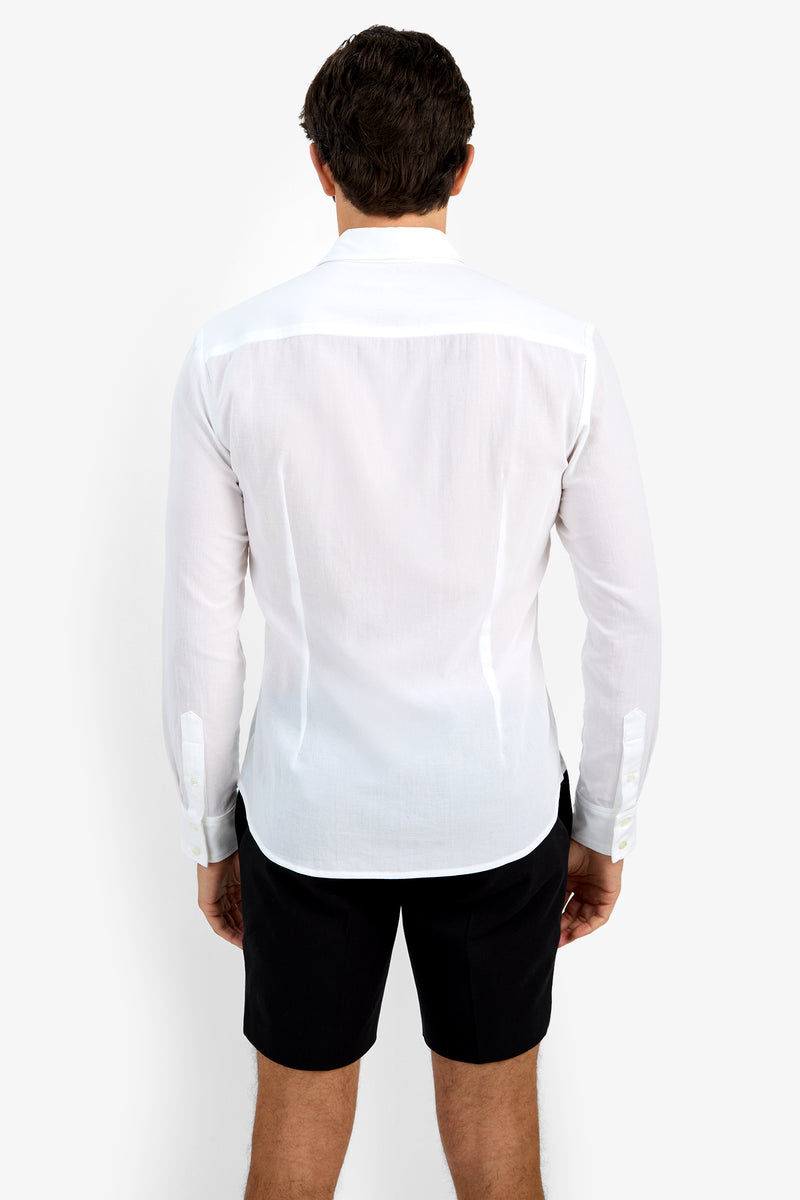 A man, with his back to the camera, wears the Aaron Shirt—a long-sleeve white dress shirt—and black shorts against a plain white background.