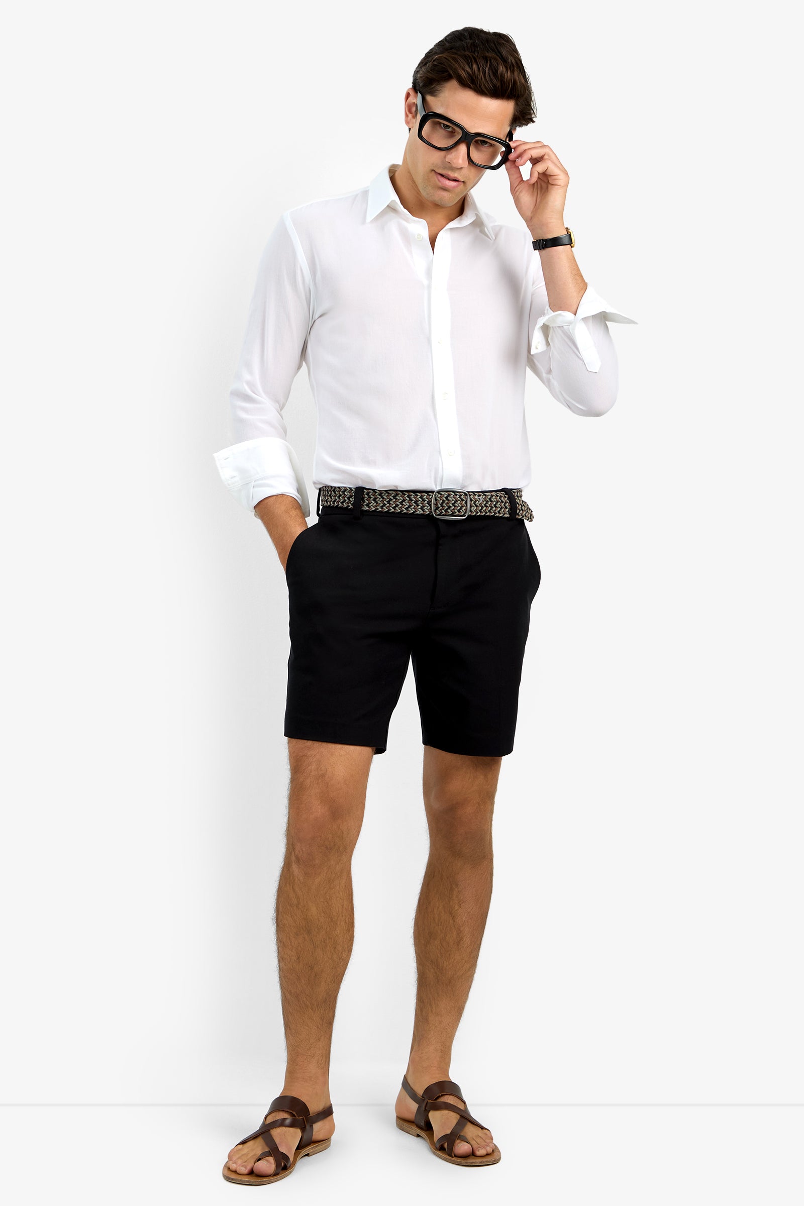 Wearing the Aaron Shirt, black shorts with a patterned belt, brown sandals, and black-framed glasses, a man stands with one hand in his pocket and the other adjusting his glasses.