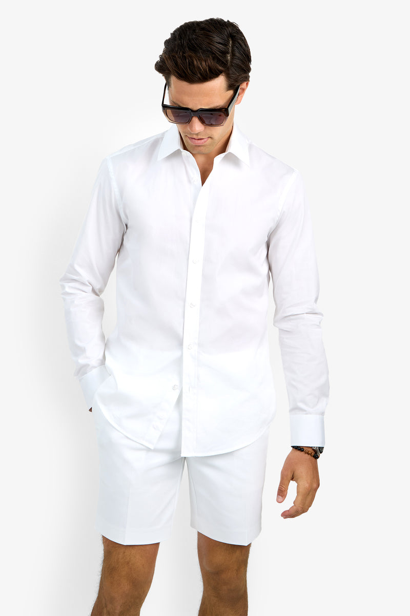 A man stands against a plain white background, looking down with one hand in his pocket. He is wearing sunglasses, white shorts, and the Aaron Shirt—a white long-sleeve button-up.