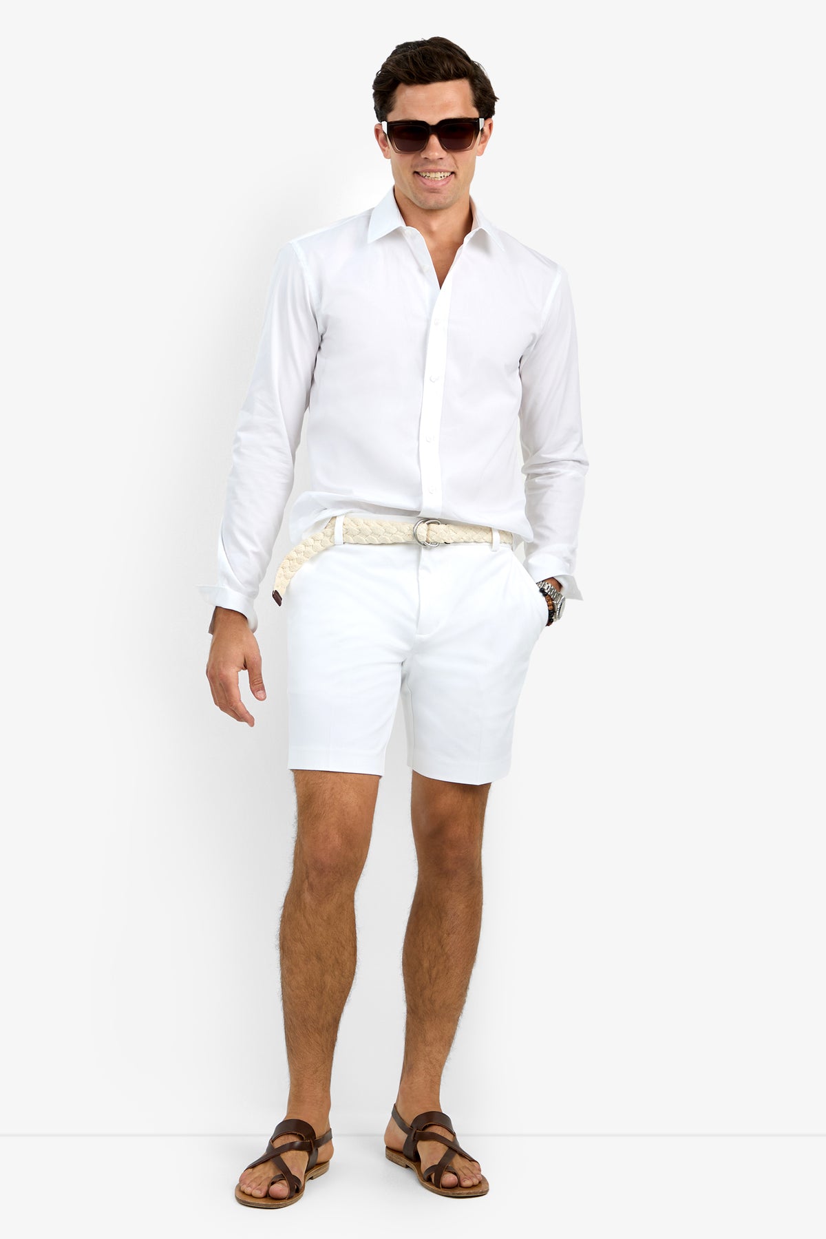 A man wearing the Aaron Shirt—a long-sleeve white button-down—paired with white shorts, brown sandals, a woven belt, and a watch stands smiling casually against a plain white background.