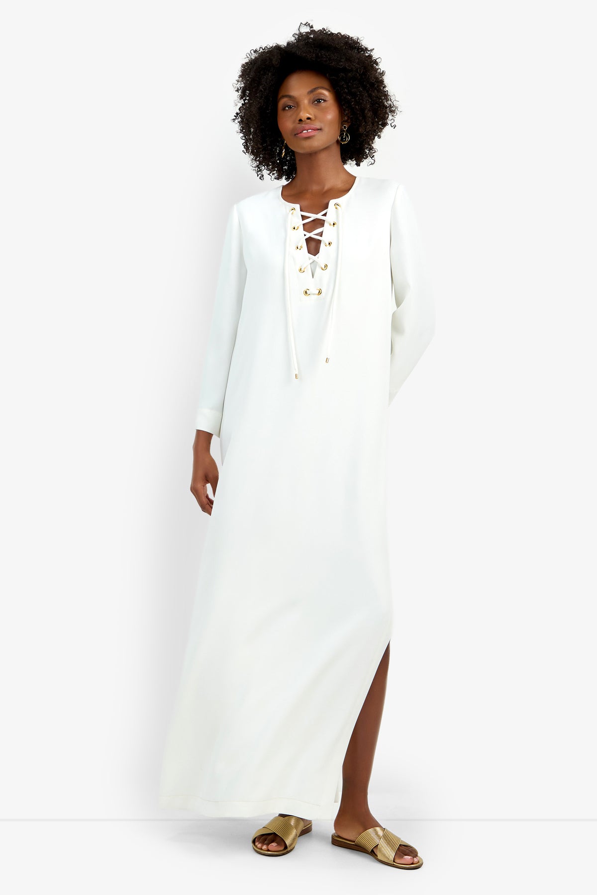 A woman with curly hair stands confidently in the Catamaran Dress—a long-sleeved white maxi with a lace-up neckline and gold eyelets—paired with gold flat sandals. The background is plain white.