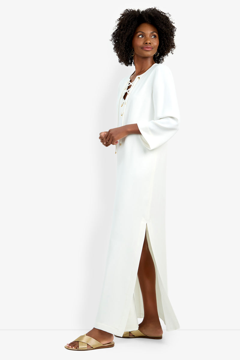 A woman in the Catamaran Dress—long, white, with a lace-up neckline and side slits—stands sideways, looking up. She has curly hair, wears gold flat sandals, and is posed against a plain white background.