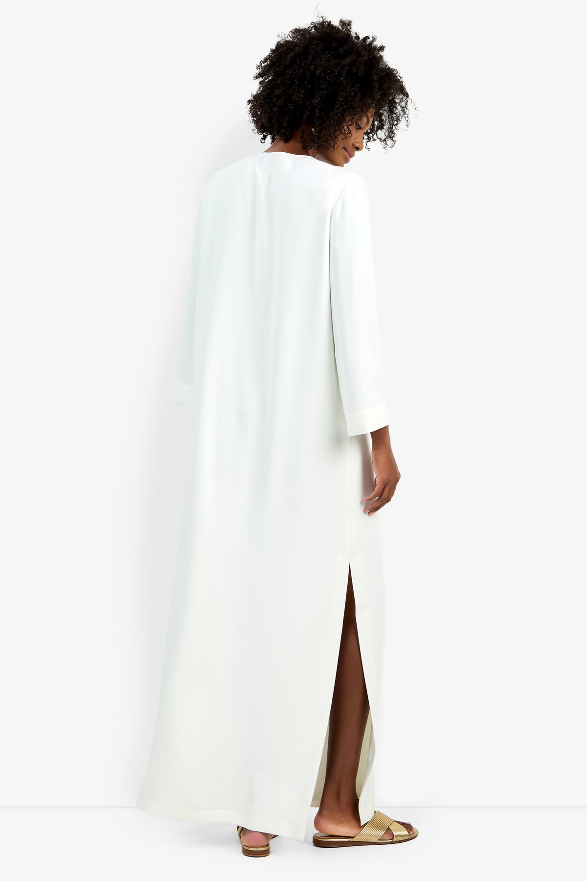 A person with curly hair stands facing away, wearing the Catamaran Dress—long, white, with three-quarter sleeves and a side slit—paired with tan slide sandals against a plain white background.