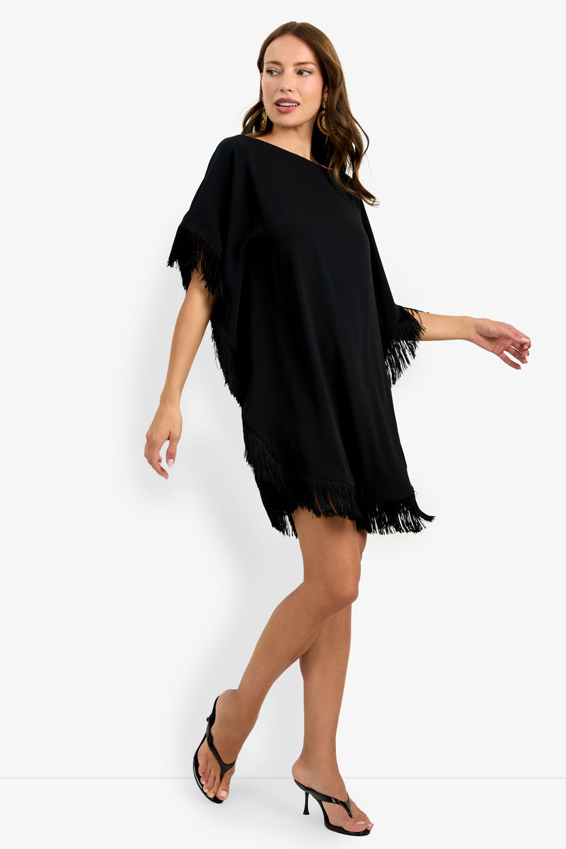 Woman in the Mini Neena Fringe Dress, a loose-fitting black mini dress with fringed edges on the sleeves and hem, wearing black heeled sandals, walks with one arm raised and looks to the side against a white background.
