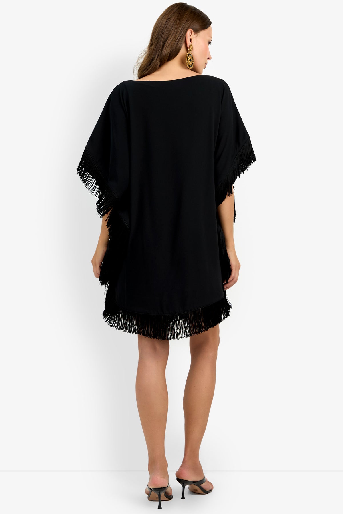 A woman stands facing away against a white background, wearing the Mini Neena Fringe Dress in black with brown hair down, black heeled sandals, and large round earrings.
