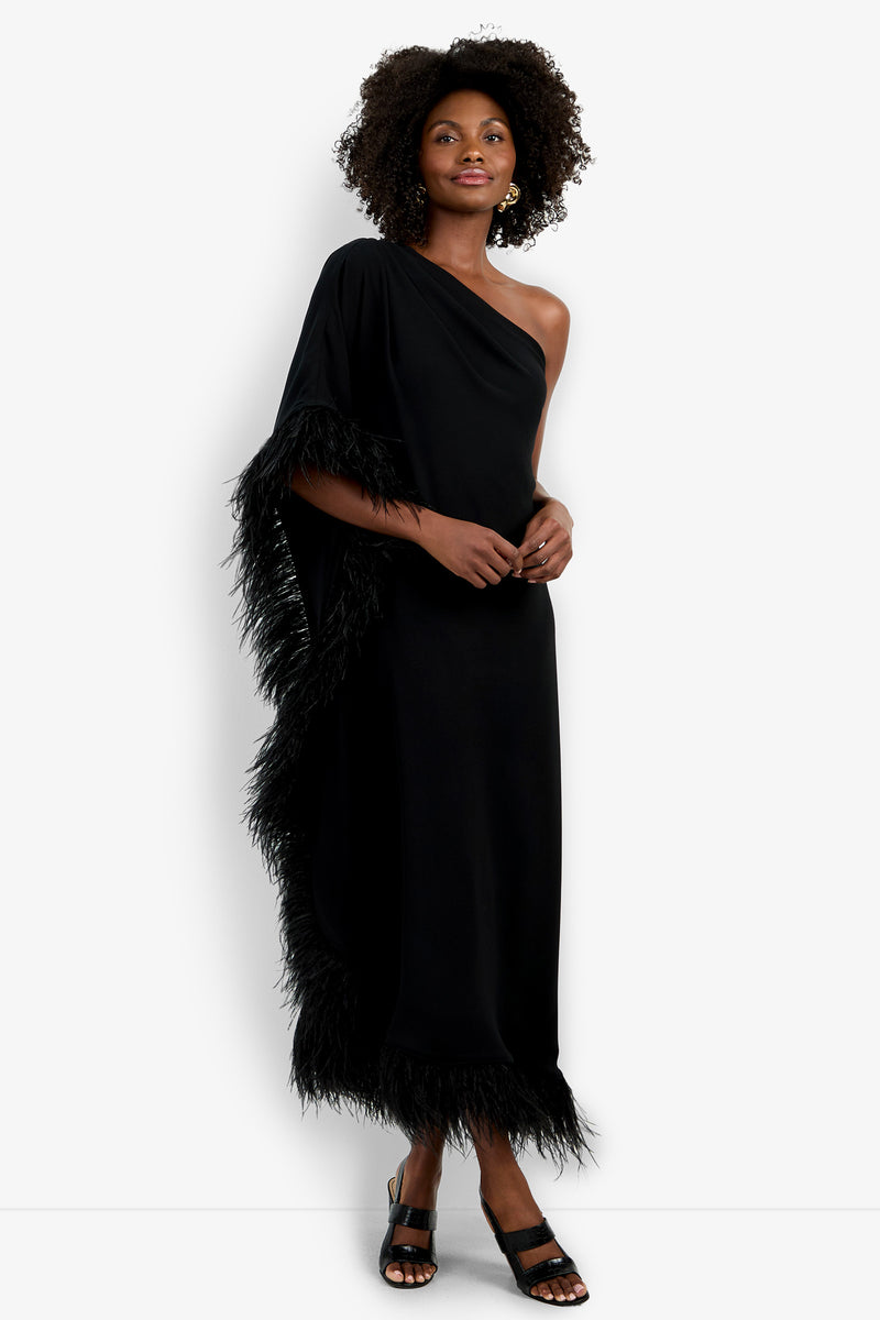 A woman smiles with hands clasped, wearing the Largo Caftan—a black, one-shoulder dress with feather-trimmed edges—paired with black sandals and gold earrings. Her naturally curly hair is styled loose.