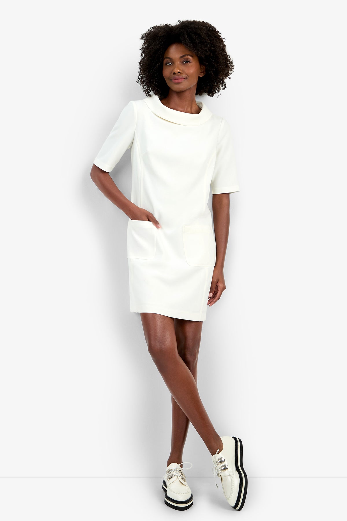 A woman with curly hair poses against a plain light background wearing the Maleko Dress—a short-sleeve white dress with two front pockets—and white platform shoes with black soles, standing with one leg crossed.
