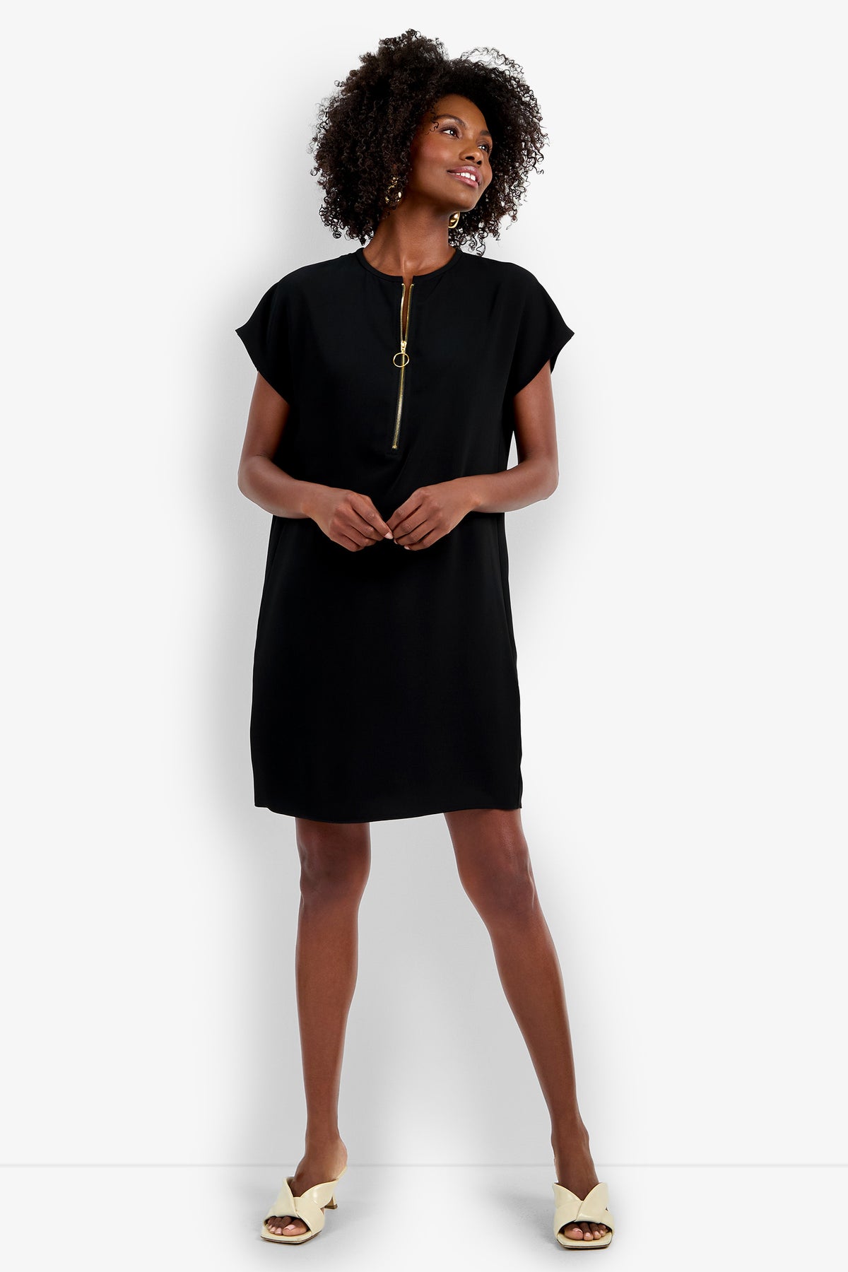 A woman smiles while wearing the Hasil Dress, a short black dress with a gold zippered neckline, paired with cream open-toe sandals, standing against a plain white background.