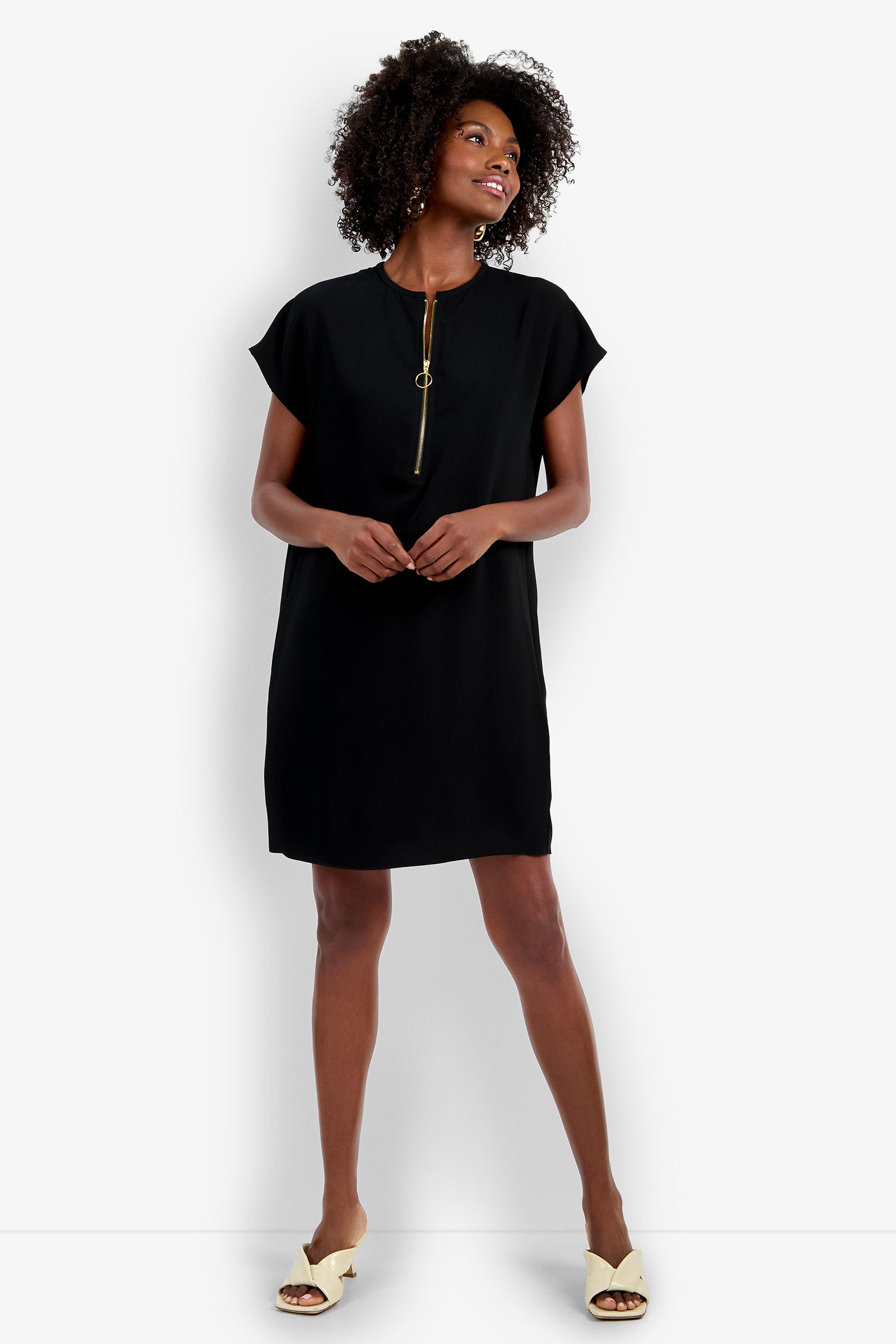 A woman smiles while wearing the Hasil Dress, a short black dress with a gold zippered neckline, paired with cream open-toe sandals, standing against a plain white background.