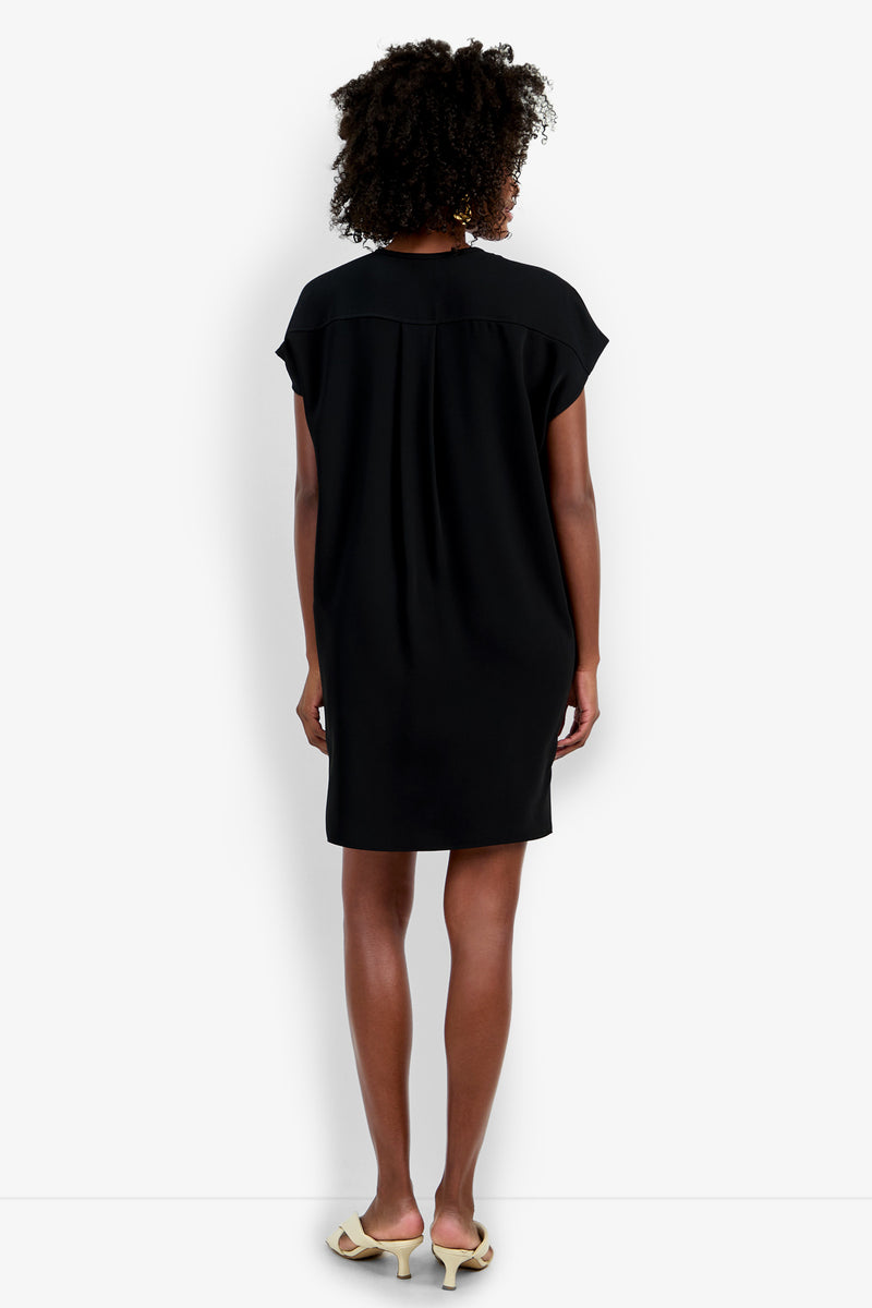 A woman faces away in the Hasil Dress, a loose-fit black short-sleeve dress with slight upper-back gathering. The dress falls above the knees and is paired with light low-heeled shoes. The background is plain white.