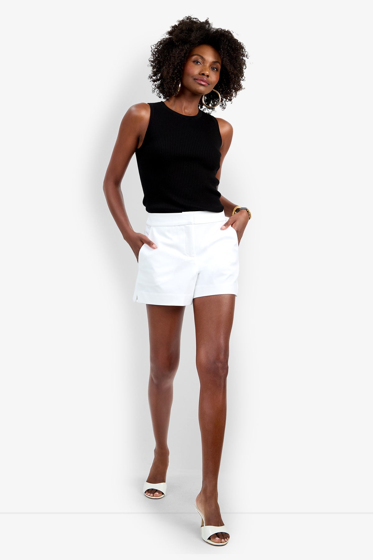 A woman stands confidently with hands in the pockets of her white Corbin Short, wearing a sleeveless black top, white heeled sandals, hoop earrings, and a bracelet. She has curly hair.