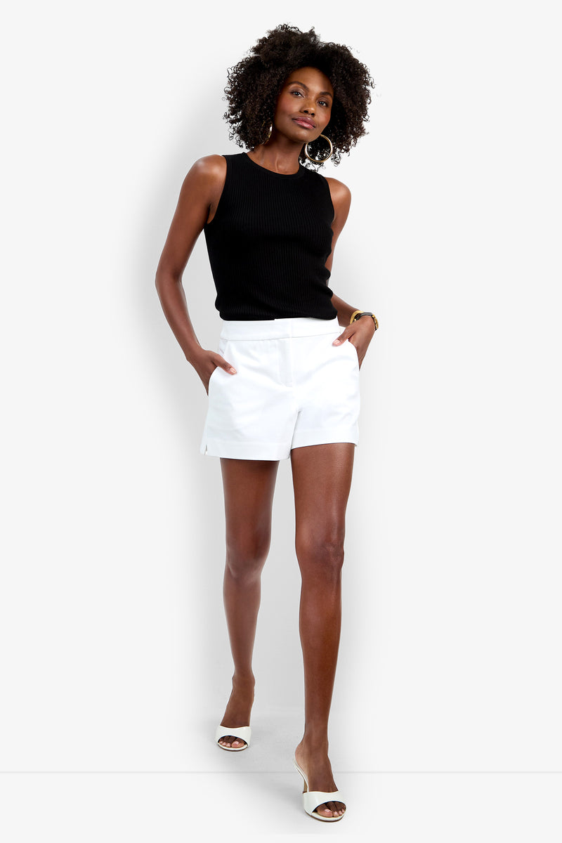 A woman stands confidently with hands in the pockets of her white Corbin Short, wearing a sleeveless black top, white heeled sandals, hoop earrings, and a bracelet. She has curly hair.