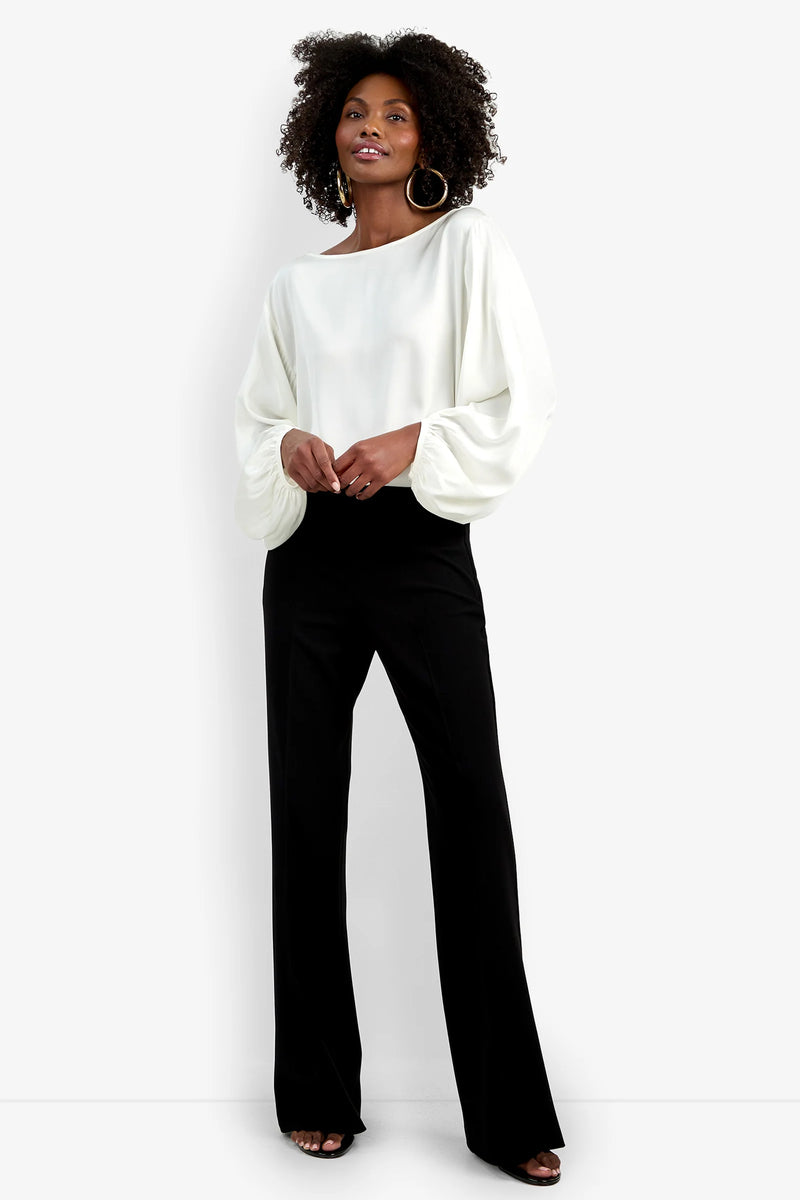 A woman stands against a white background wearing the Chimayo Pant—black, wide-leg pants—paired with a white long-sleeve blouse with billowy sleeves, hoop earrings, and open-toe black shoes.