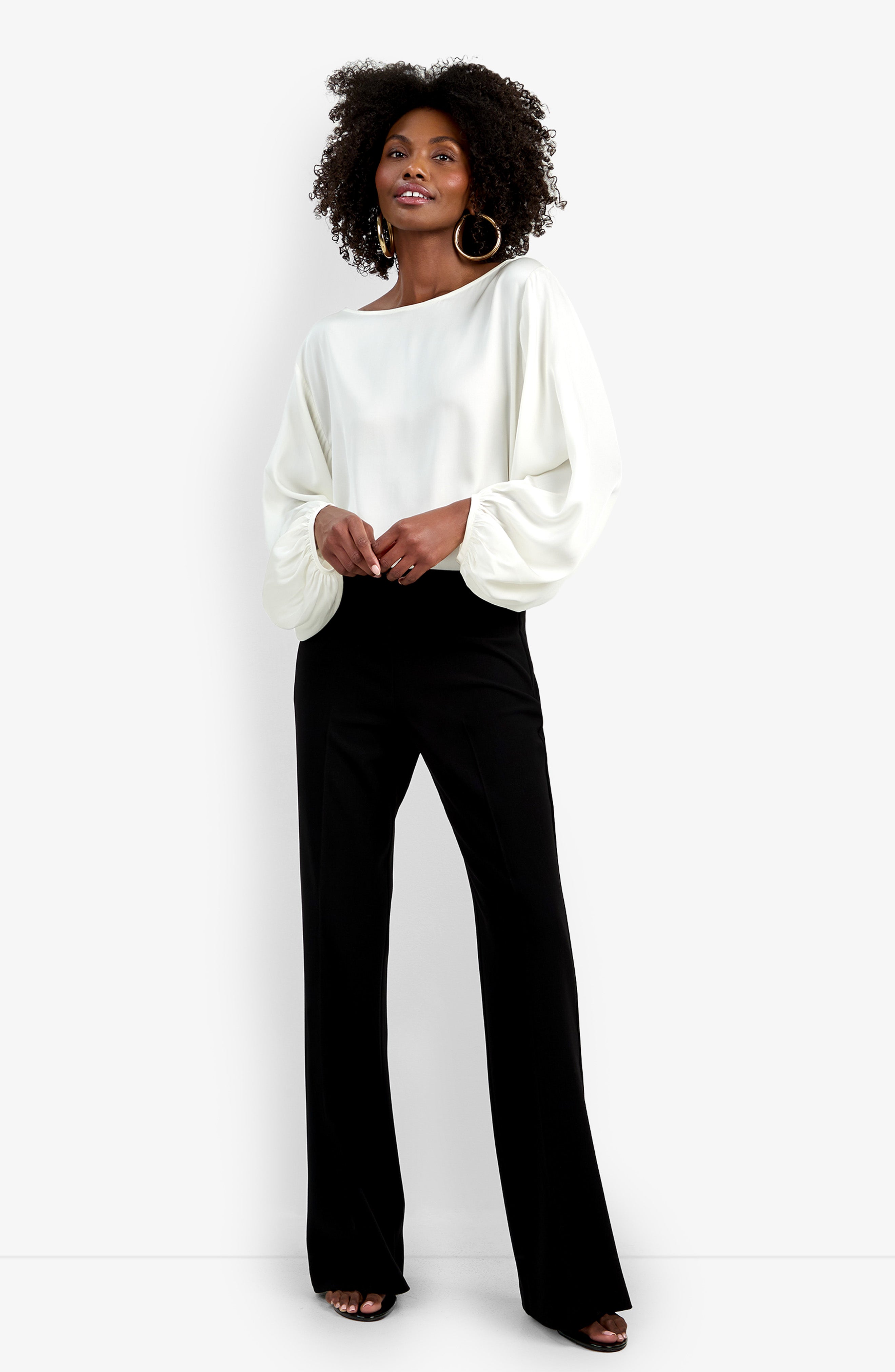 A woman smiles against a white background, wearing black Chimayo Pant wide-leg trousers, a loose white long-sleeve top with billowy sleeves, hoop earrings, and open-toed shoes. She stands with her hands lightly clasped.