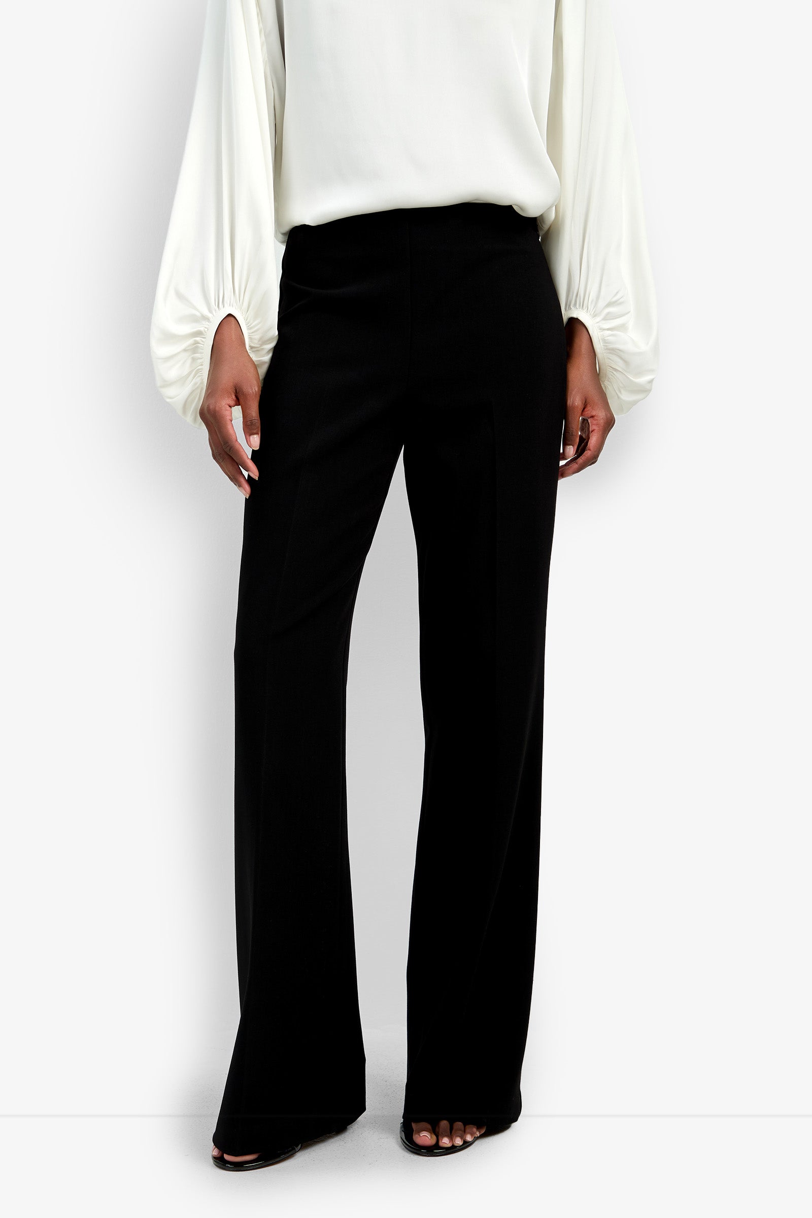 A person wearing high-waisted, wide-leg black Chimayo Pants and a white long-sleeve blouse with billowy sleeves, standing against a plain white background.