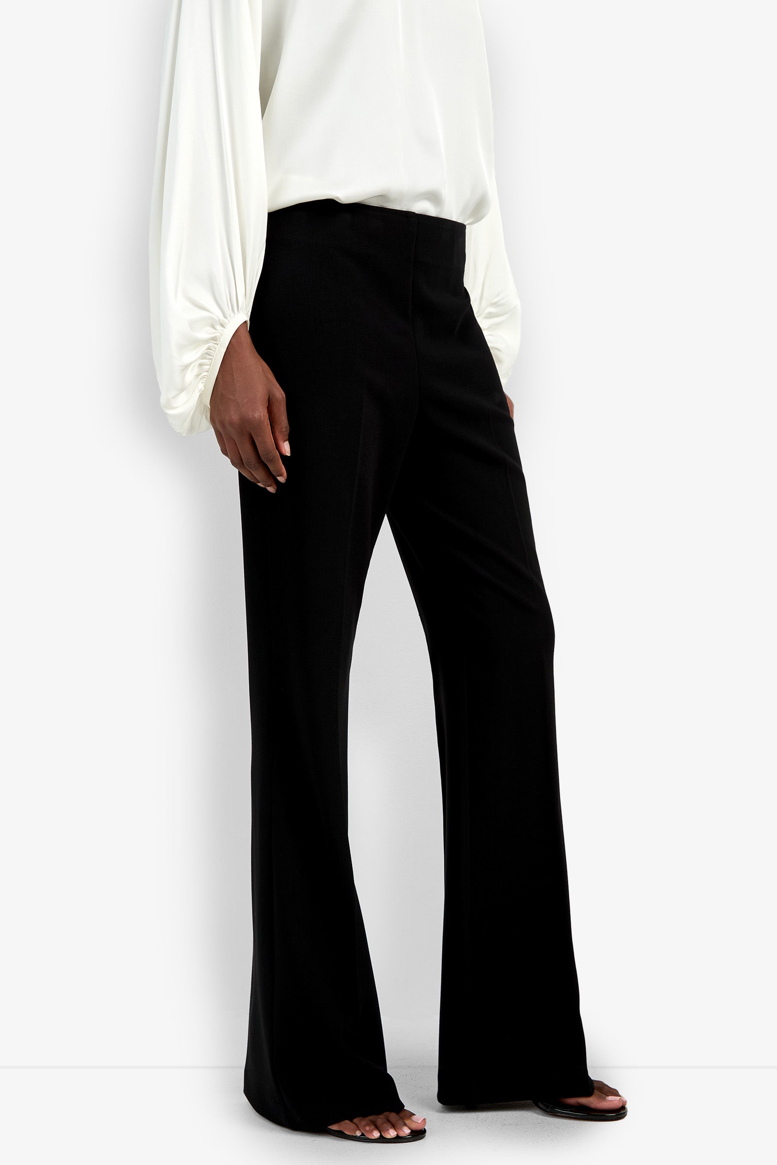 A person is seen from the shoulders down wearing high-waisted, black Chimayo Pants and a white long-sleeve blouse, set against a white background.