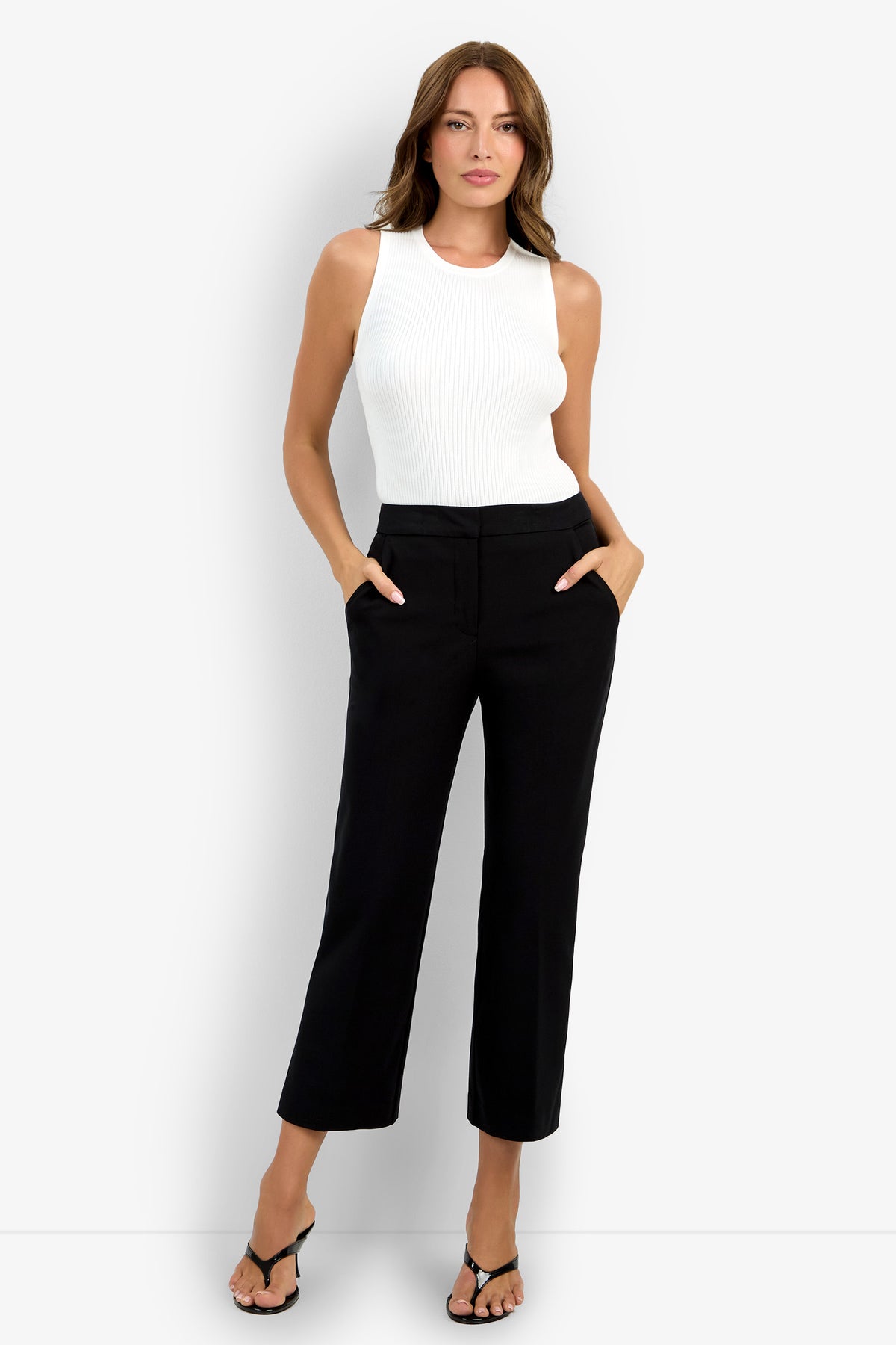 A woman stands against a white background wearing black Lulu Pant wide-leg trousers, a sleeveless white ribbed top, and black heeled sandals, with her hands in her pockets.