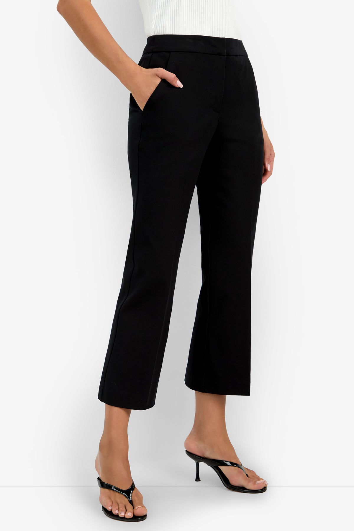 A person stands with one hand in their pocket against a white background, wearing black Lulu Pants (cropped, straight-leg), a white top, and black heeled sandals.