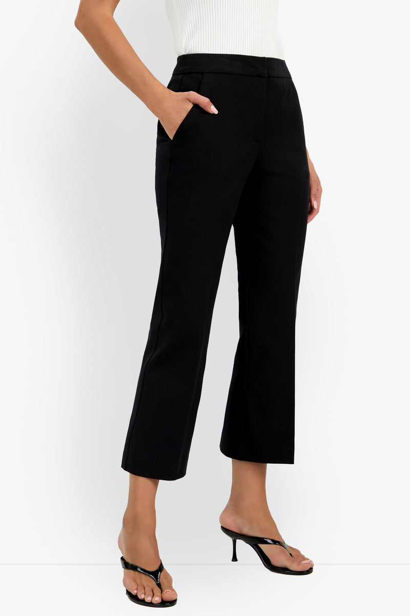A person wearing the black Lulu Pant, styled with a white sleeveless top and black heeled sandals, stands with one hand in their pocket against a plain white background.