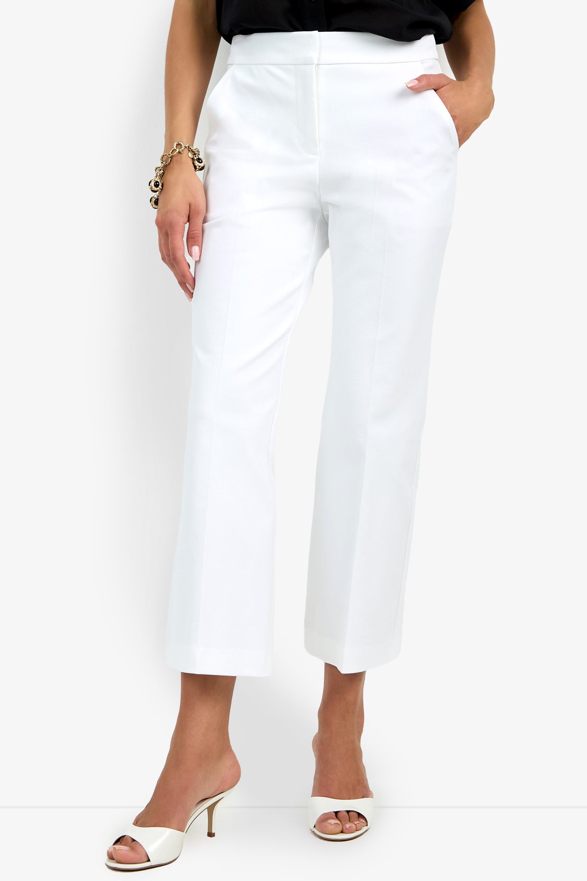 Woman wearing the Lulu Pant—white, high-waisted, ankle-length trousers with a straight cut—paired with a black top, white open-toe heels, and a chunky bracelet; her hand is in one pocket.