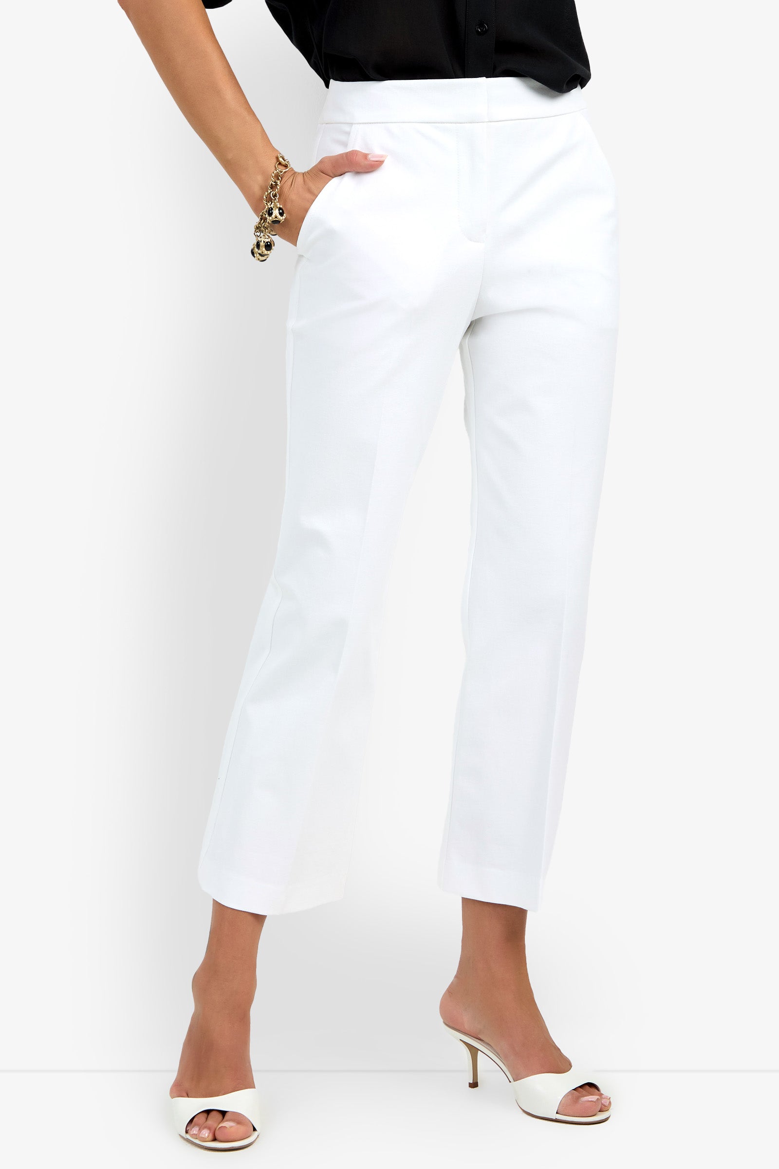 A person wearing the Lulu Pant in white with a black top, white heeled sandals, and a gold bracelet, standing with one hand in their pocket against a plain background.