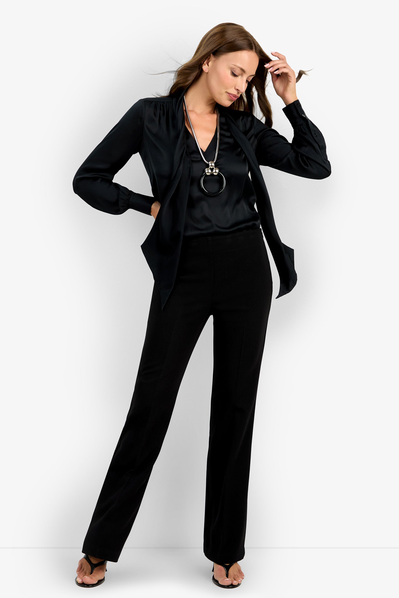 A woman stands against a white background wearing the Chimayo Pant, a black blouse, open-toed heels, and a long pendant necklace, with one hand touching her hair and the other by her side.