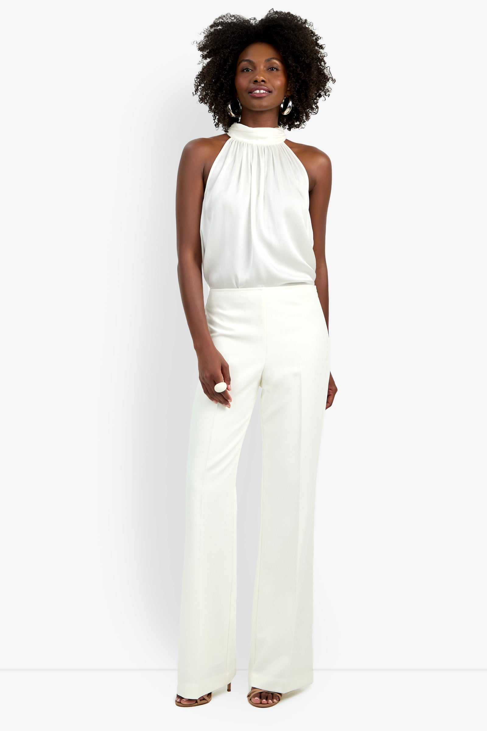 A woman with curly hair smiles, wearing the CHIMAYO 2 PANT—high-waisted, wide-leg trousers made from soft polyester and elastane—with a sleeveless white halter top and open-toed heels, posing against a plain white background.