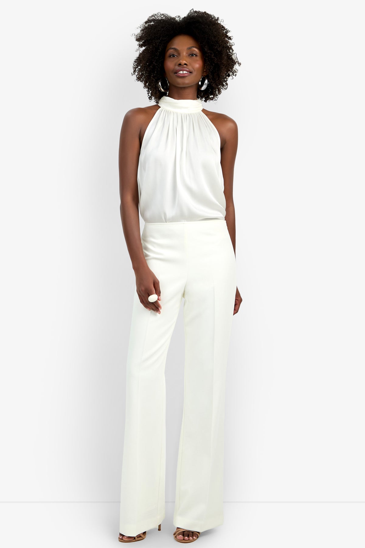 A woman with curly hair smiles confidently against a white background, wearing the Chimayo Pant—high-waisted, wide-leg white trousers—paired with a sleeveless high-neck blouse and open-toe heels.
