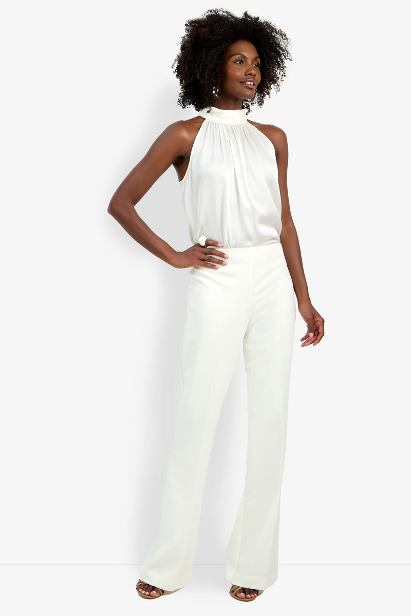 A woman stands against a plain background, posing with one hand on her hip, dressed in high-waisted, wide-leg CHIMAYO 2 PANTs paired with a sleeveless white halter-neck blouse and brown sandals.