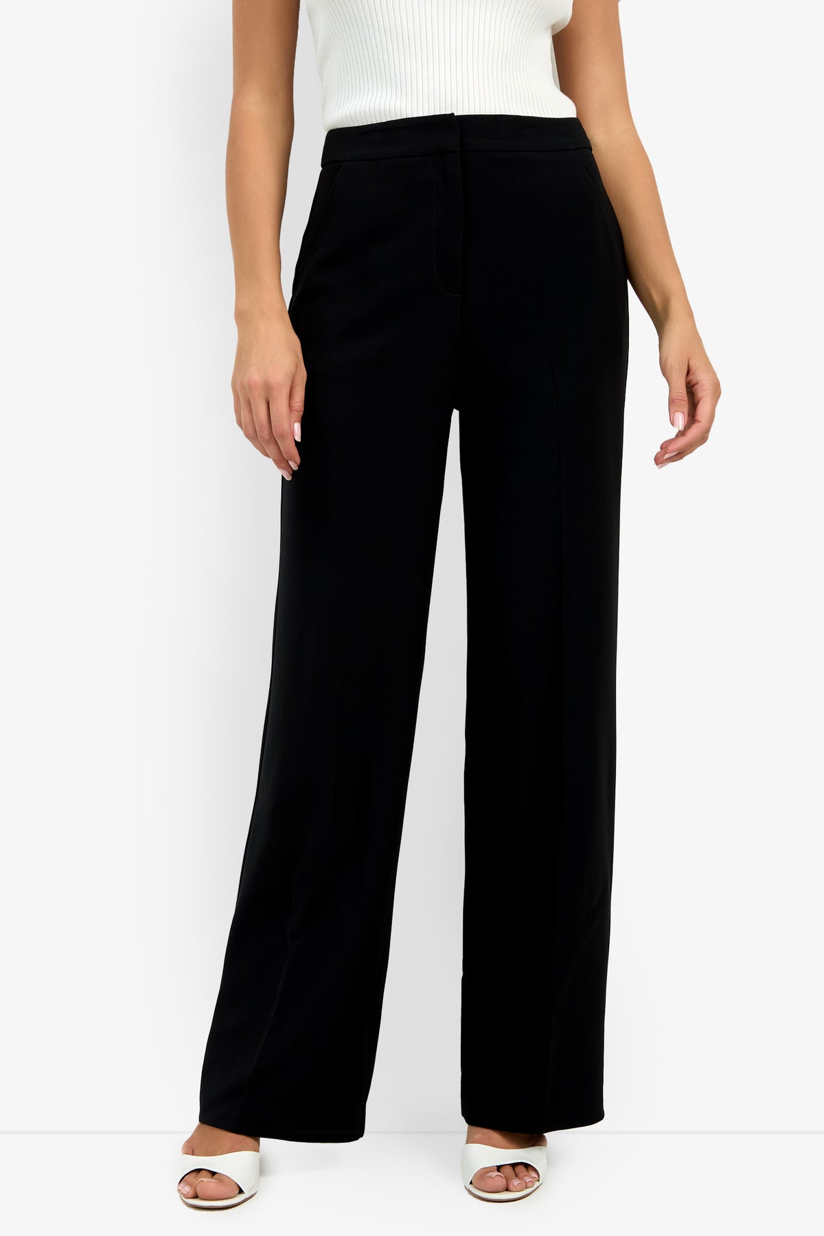 A person models the Sadie Pant in black with a white sleeveless ribbed top and white open-toe heels against a plain white background. The image is cropped at the shoulders.